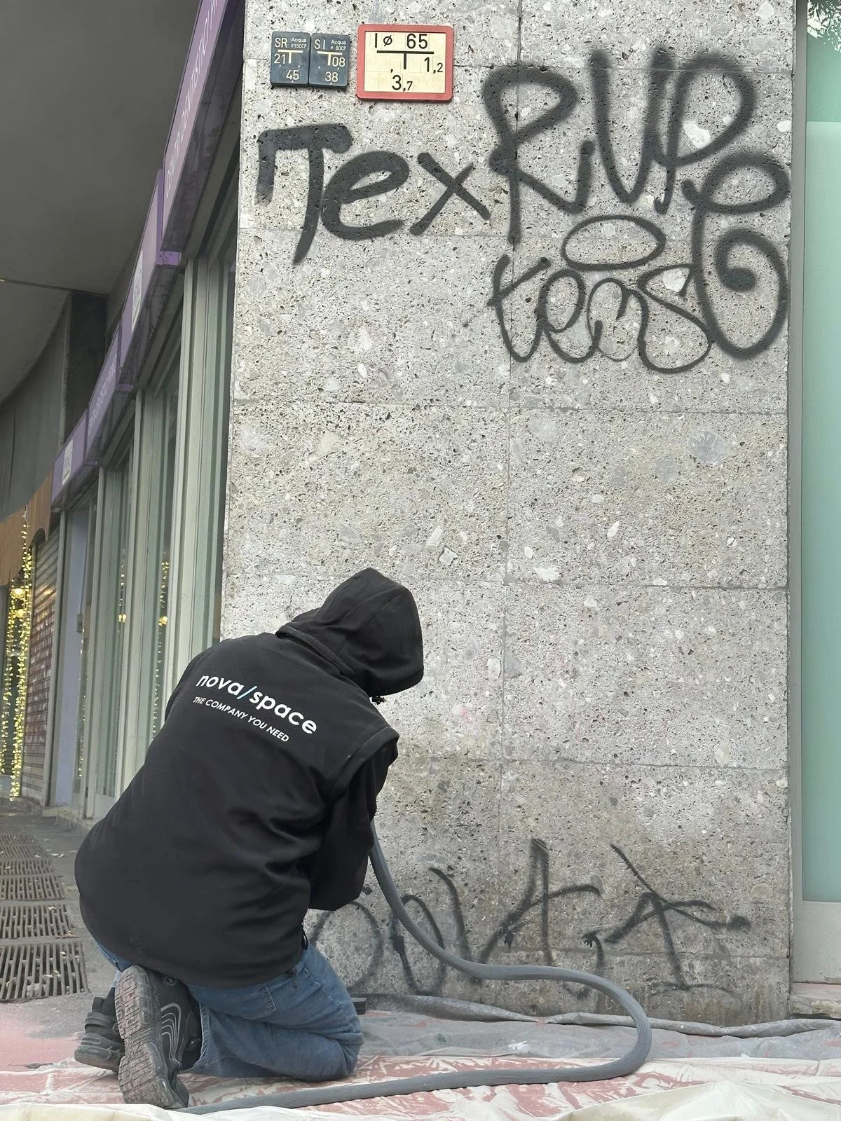 Persona in ginocchio mentre lavora con un tubo nero attaccato alla parete di un edificio di pietra, con graffiti e segnali stradali sopra.