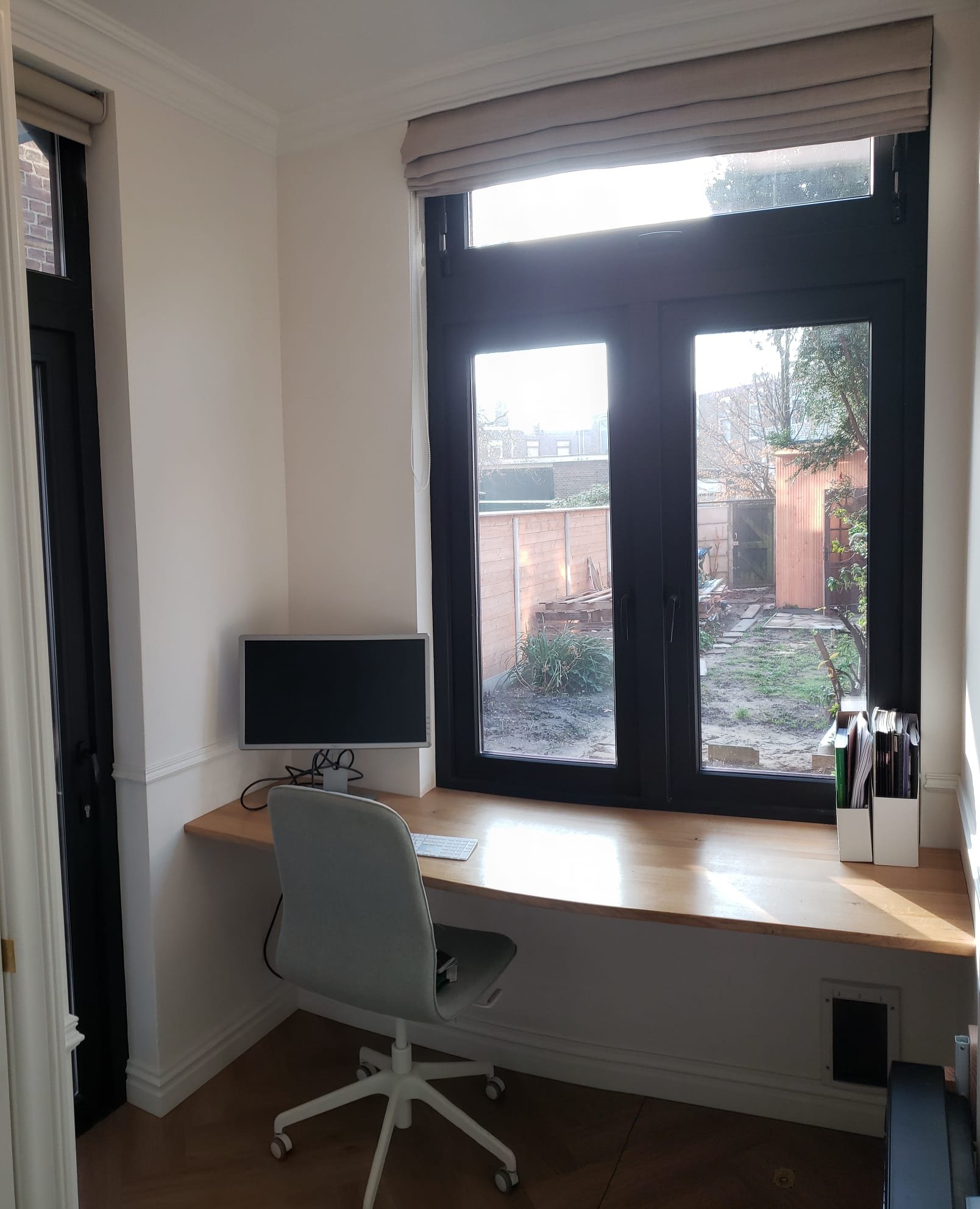 Home office space with a wooden desk positioned in front of large windows, a computer monitor and keyboard on the desk, an office chair, and a small collection of files or notebooks stored in a white holder.