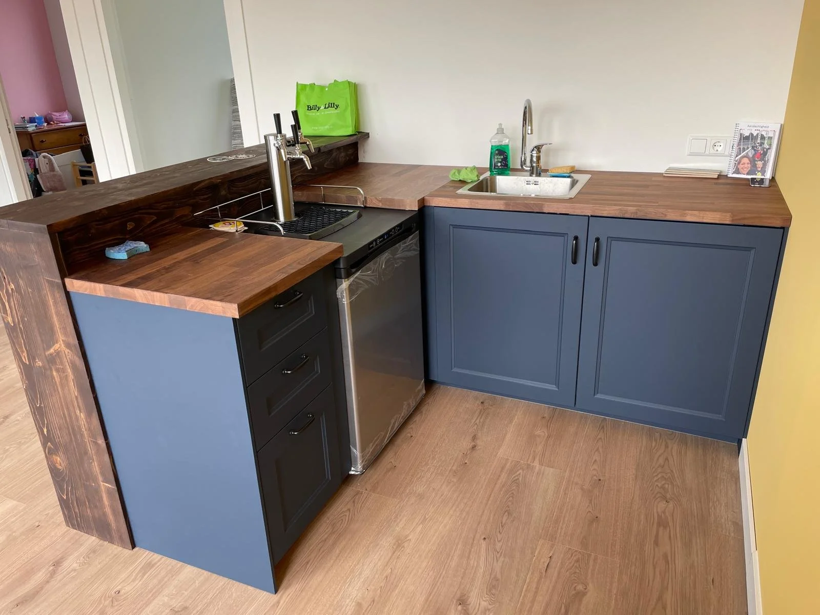 Small kitchen with wooden countertop, blue cabinet doors, and a small stainless steel dishwasher. There is a sink with a green dish soap bottle and a green sponge. On the countertop are a framed photo, a green shopping bag, and some coasters. A wall outlet and an electrical switch are visible on the wall.