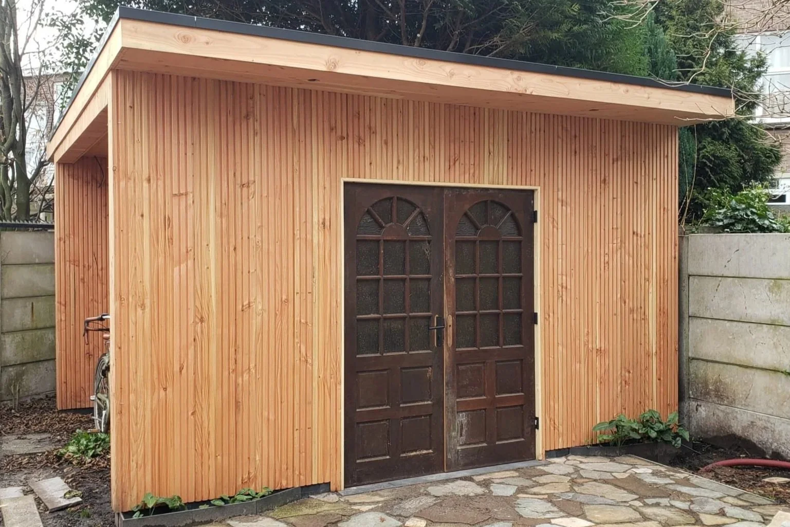 Small wooden shed with double arched glass doors, located in a backyard with a stone path in front and concrete walls on either side.