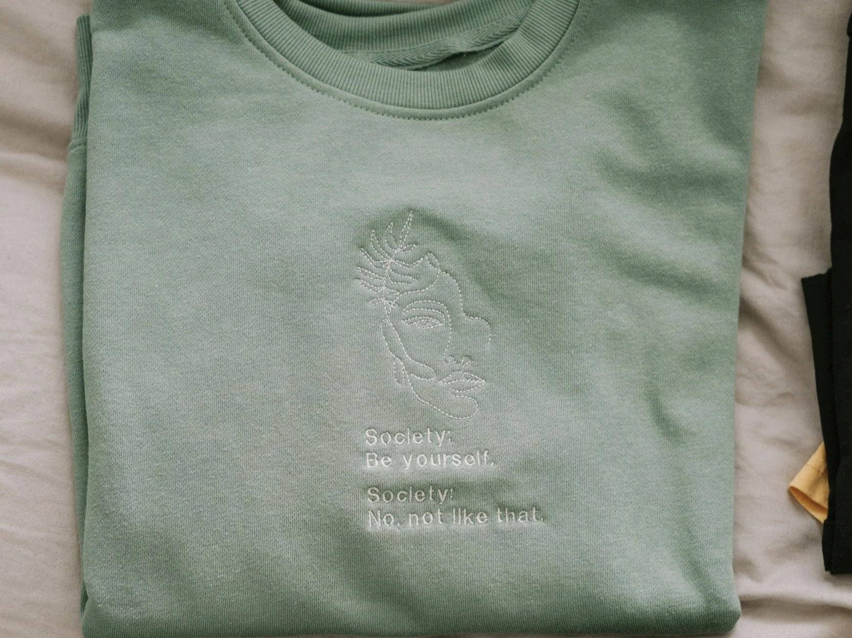 Light green sweatshirt with white embroidered face and text that reads: Society: Be yourself. Society: No, not like that.