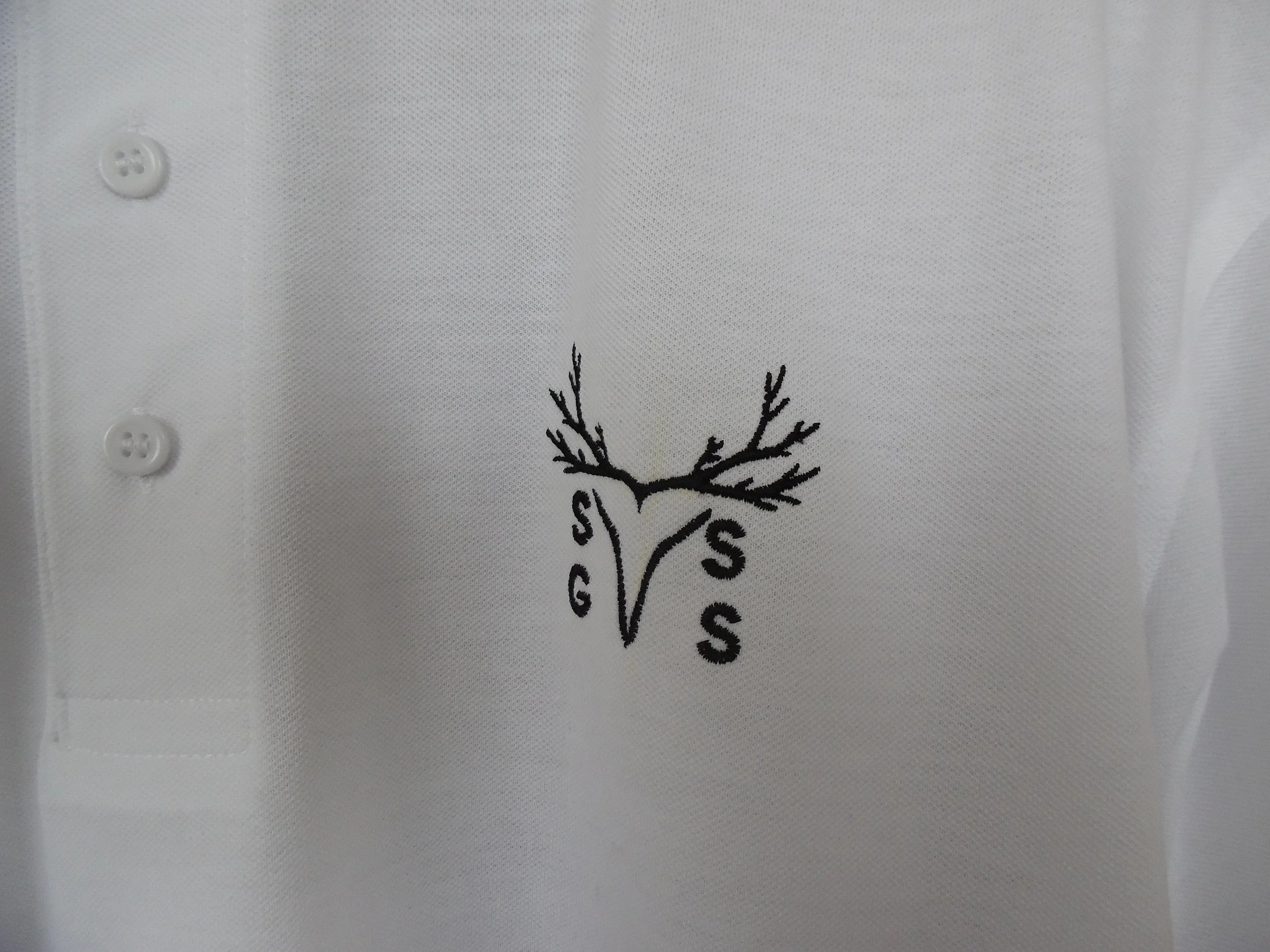 Close-up of a white polo shirt with a black embroidered logo of a deer head with antlers and the letters 'G', 'S', 'S' surrounding it.
