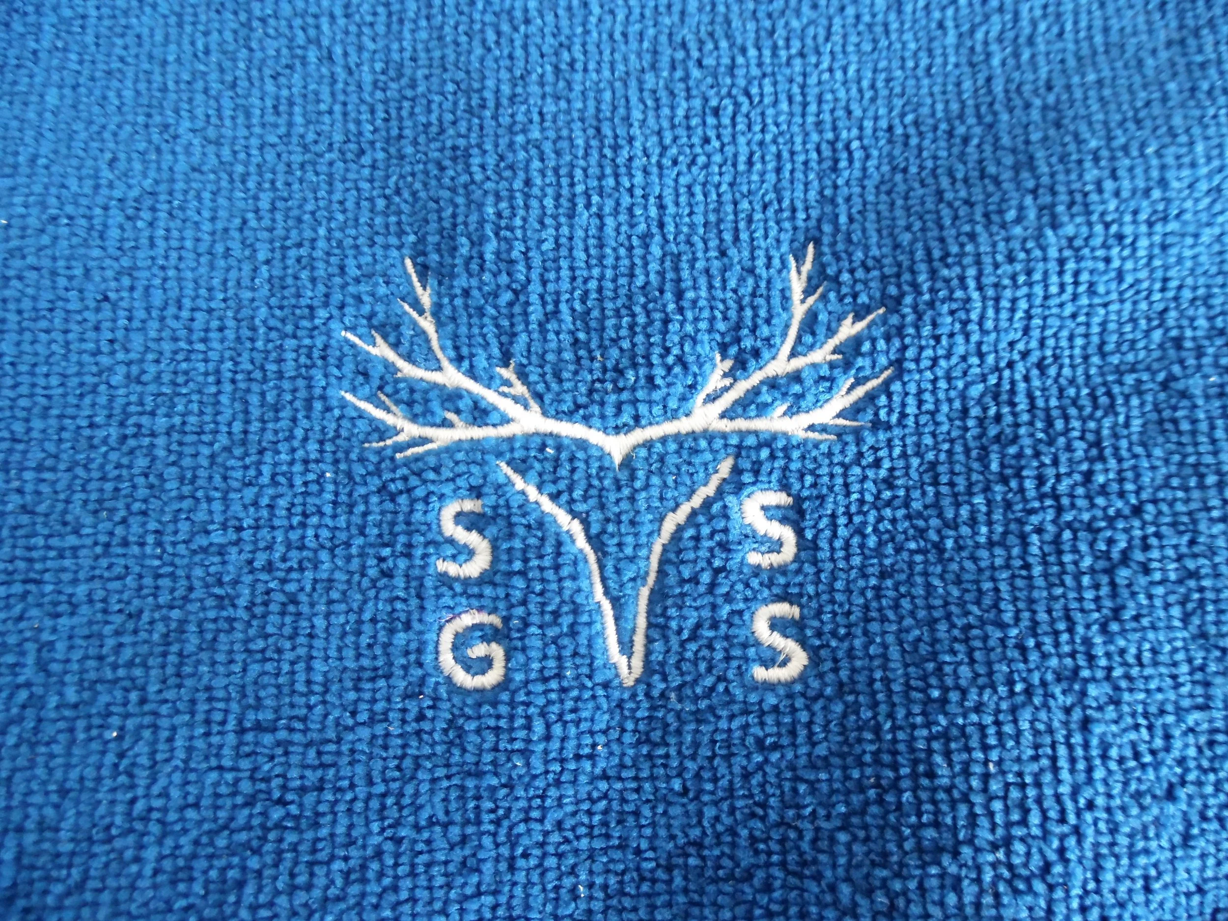 Embroidery of a stylized deer head with antlers on blue fabric, with the letters S, G, and S surrounding the design.