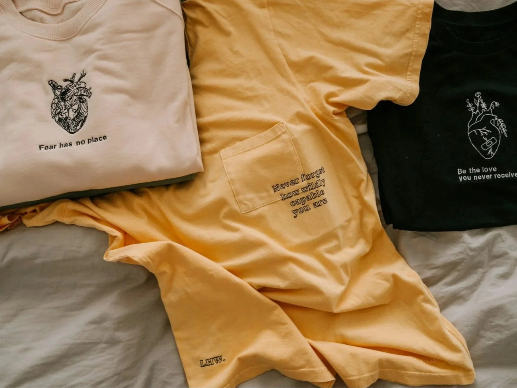 Three T-shirts with printed designs and messages, one white with a heart illustration and text, one yellow with a pocket and embroidered message, and one black with a line drawing and text, all laid out on a bed.