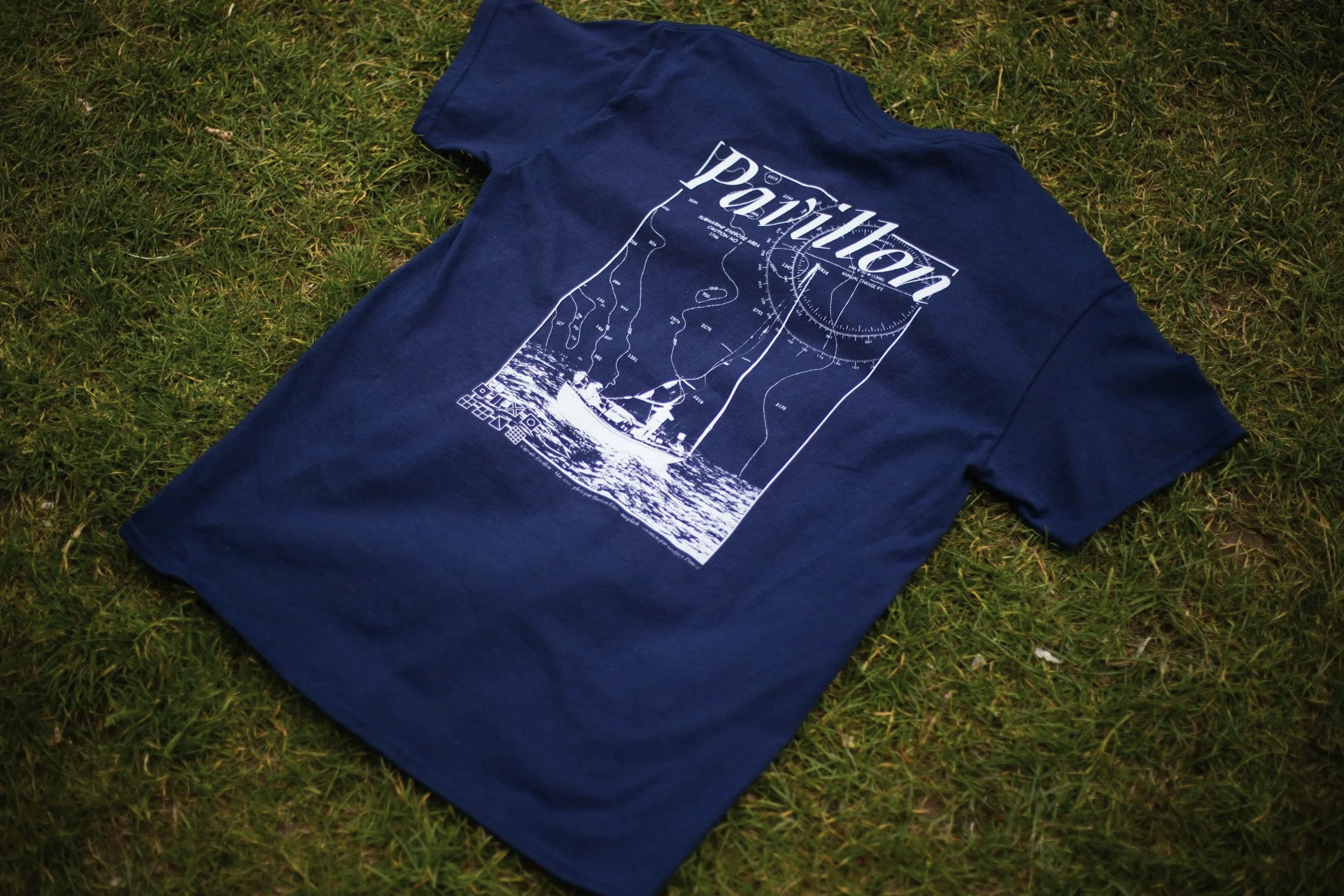 A navy blue T-shirt with a white graphic of a map, compass, and a sailboat, laid flat on grass.