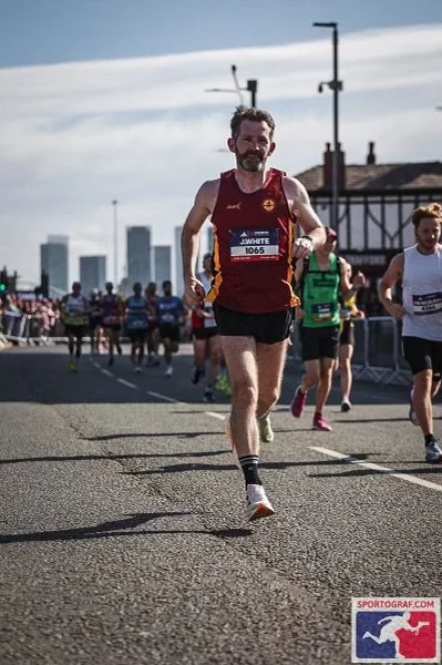 A man running in a marathon or race, wearing a maroon sleeveless shirt with a race bib numbered 1065, and black shorts, with other runners and a city skyline in the background.