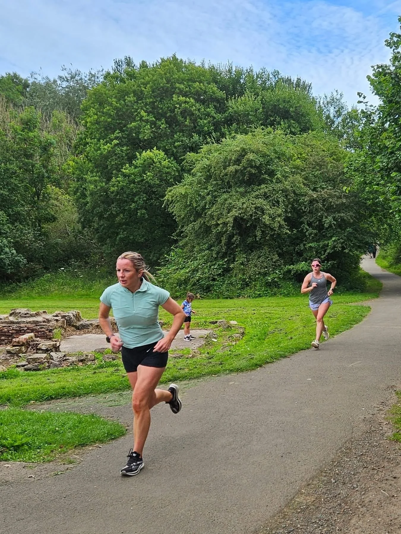 Two women and a child jogging on a paved trail in a park with green trees and bushes.