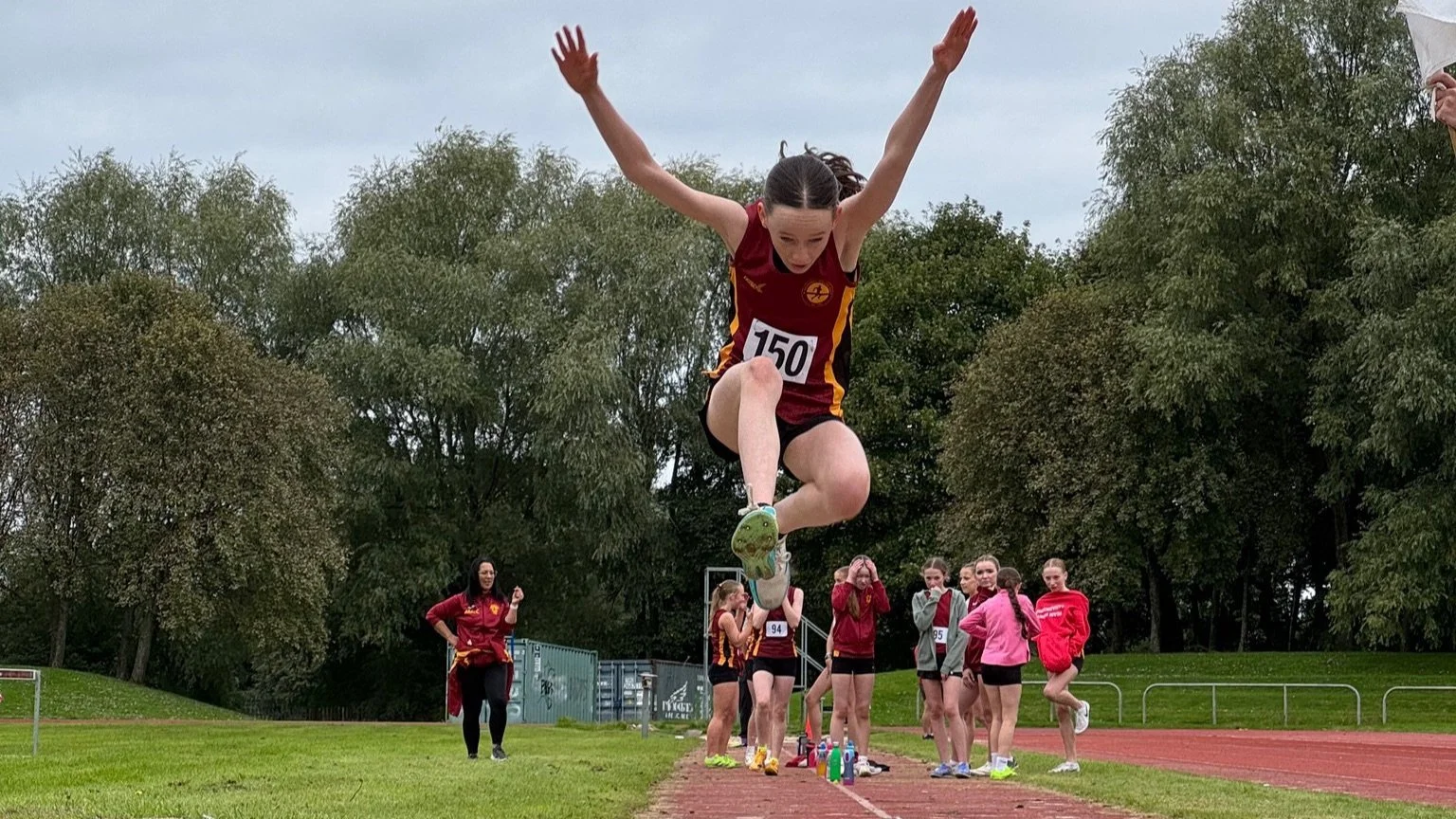 A female athlete is captured mid-air as she jumps over a hurdle during an outdoor track and field event. She is wearing a maroon and yellow uniform with the number 150.