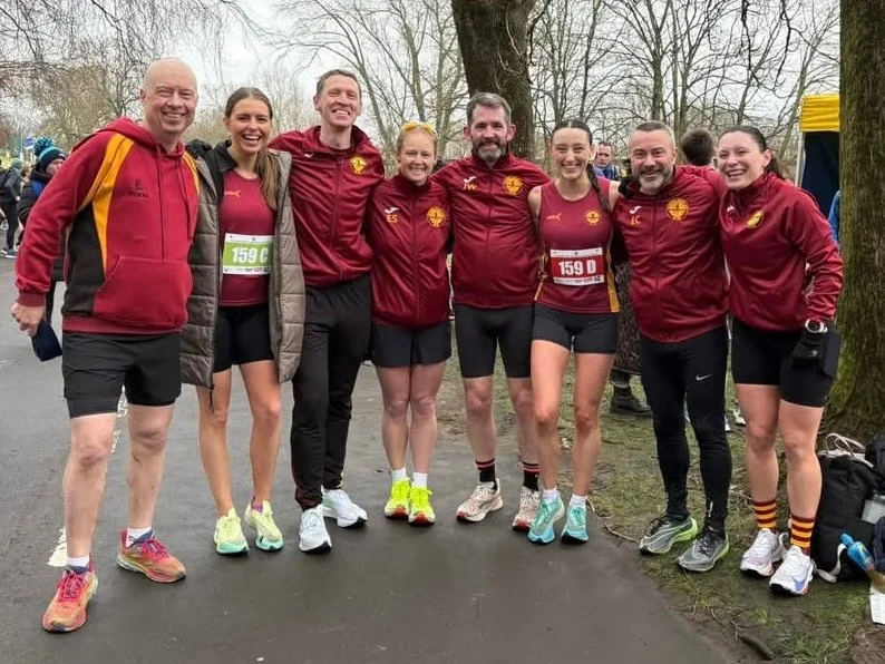 A group of eight runners in matching maroon and yellow athletic clothing, standing outdoors on a paved path, smiling with trees in the background.