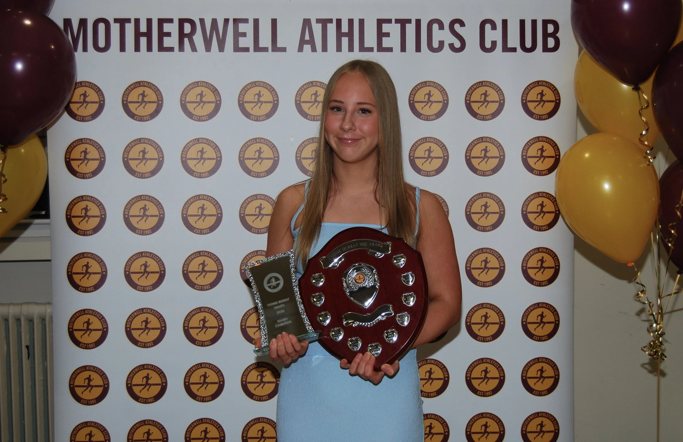 A young woman in a light blue dress holding an award plaque in front of a backdrop with the logo of Motherwell Athletics Club and balloons on either side.