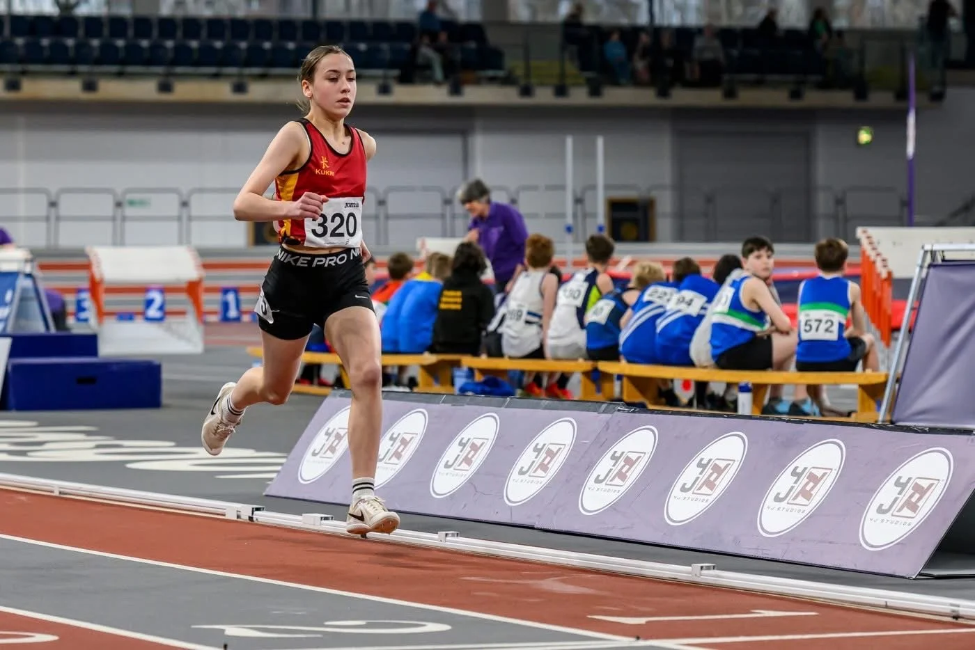 A female athlete running on an indoor track during a race, wearing a red and black sports outfit with number 320, with other athletes sitting on benches in the background.