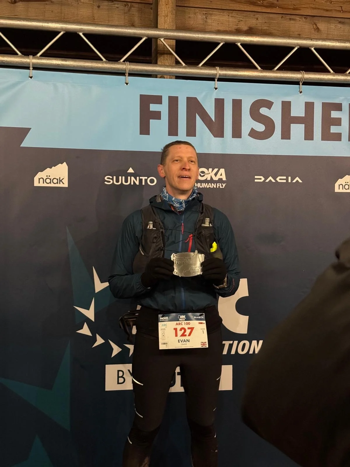 Male runner at the finish line holding a finisher medal, wearing a bib with the number 127, dressed in athletic gear, in front of a backdrop labeled 'FINISH' with sponsor logos.