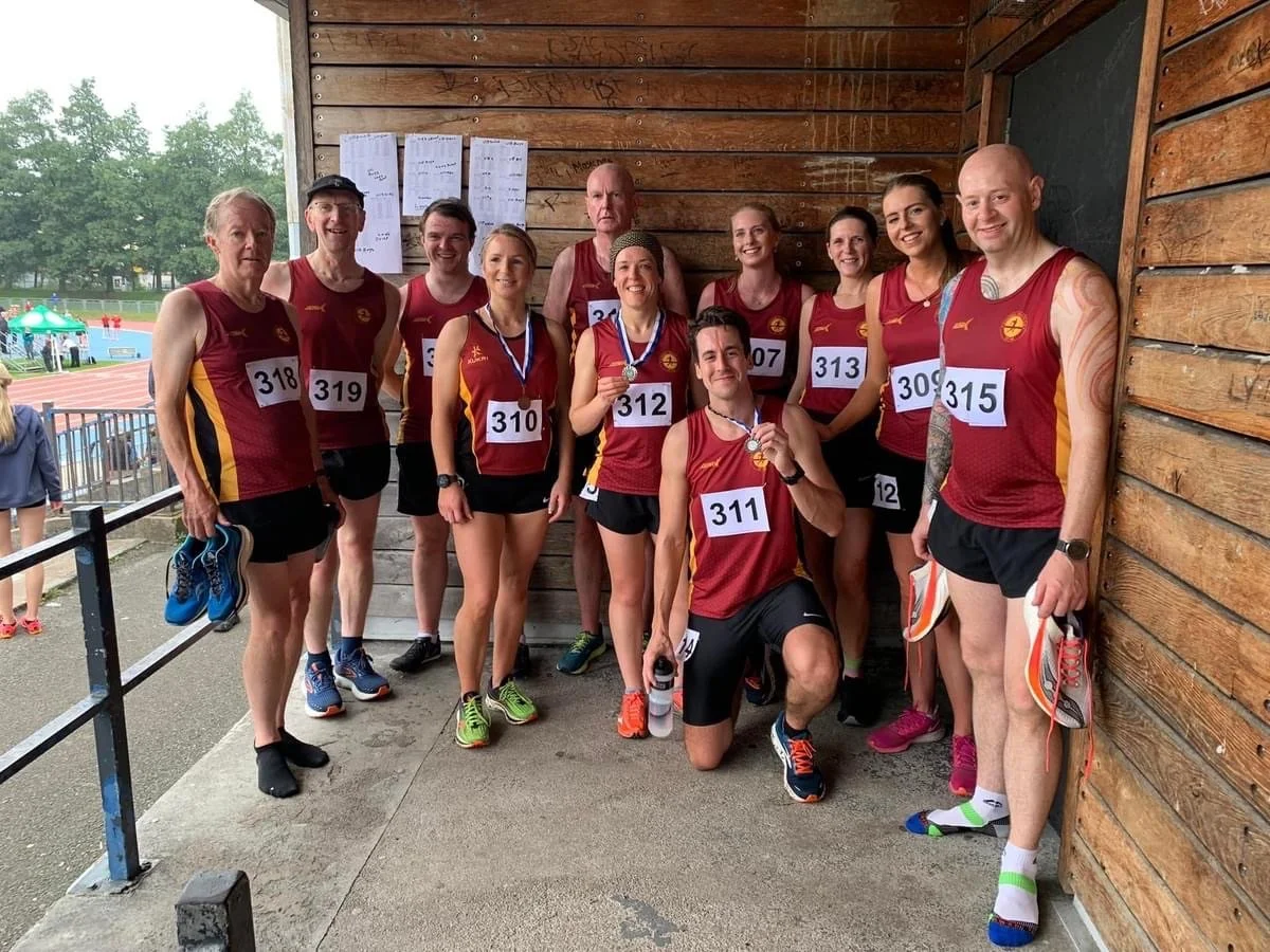 Group of athletes in matching maroon and yellow uniforms with race numbers, standing together after a race, some wearing medals, indoors against a wooden wall, with a track and field in the background.