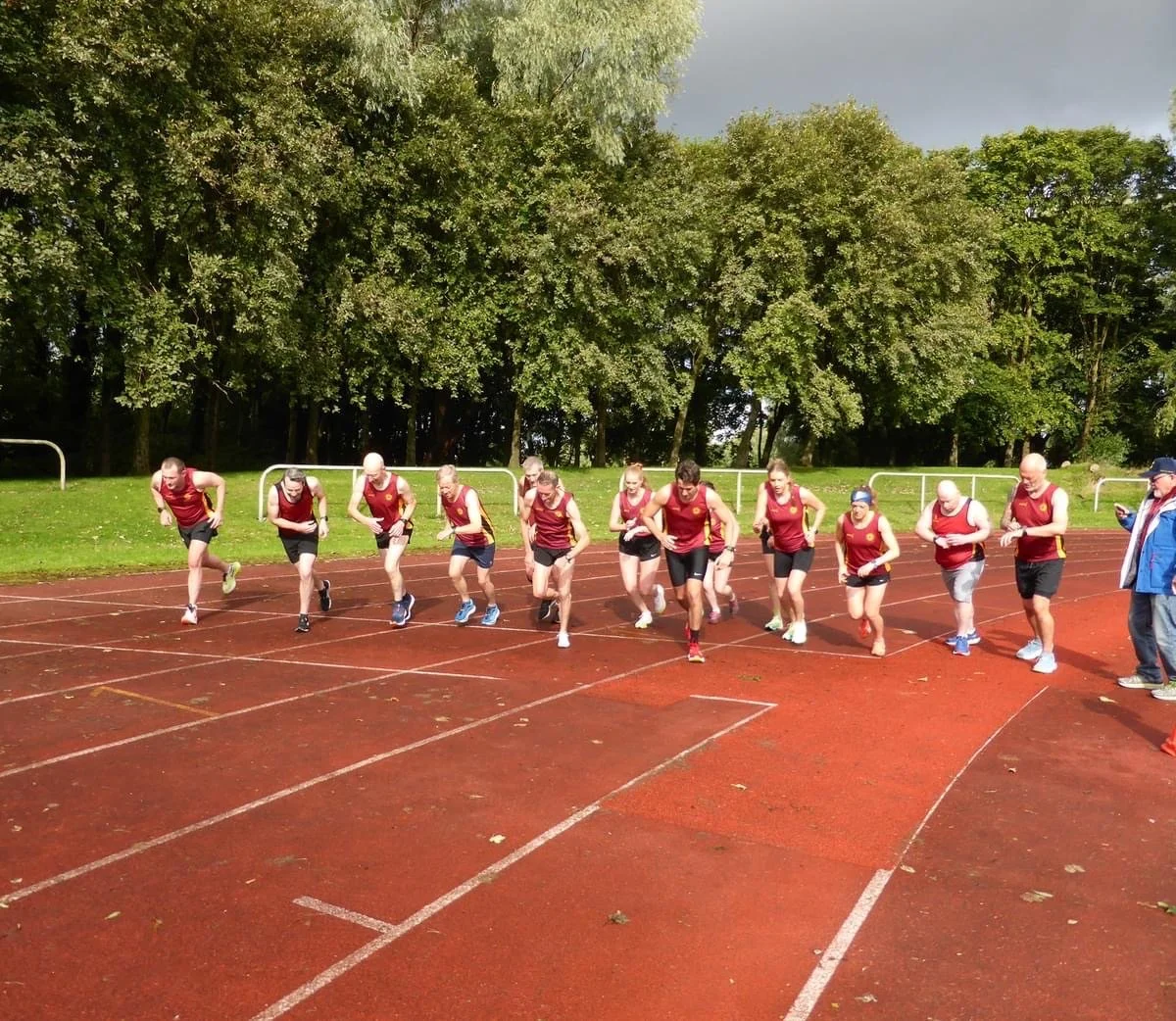 A group of runners starting a race on a red track, dressed in matching maroon athletic jerseys and shorts, with some wearing sunglasses and headbands, outdoors on a cloudy day with green trees in the background.