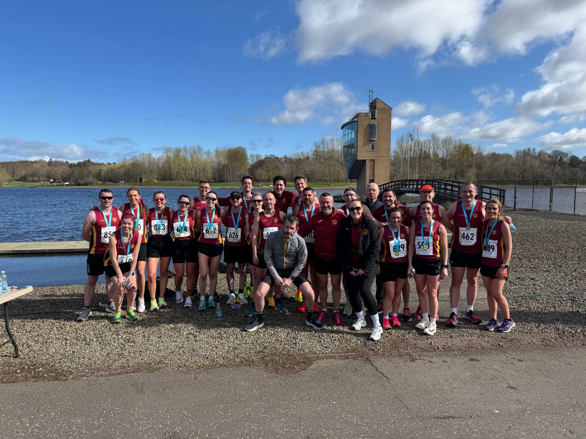 A group of marathon runners wearing matching red and black athletic uniforms and medals, posing for a photo outdoors near a lake with a bridge and a modern building in the background on a sunny day.