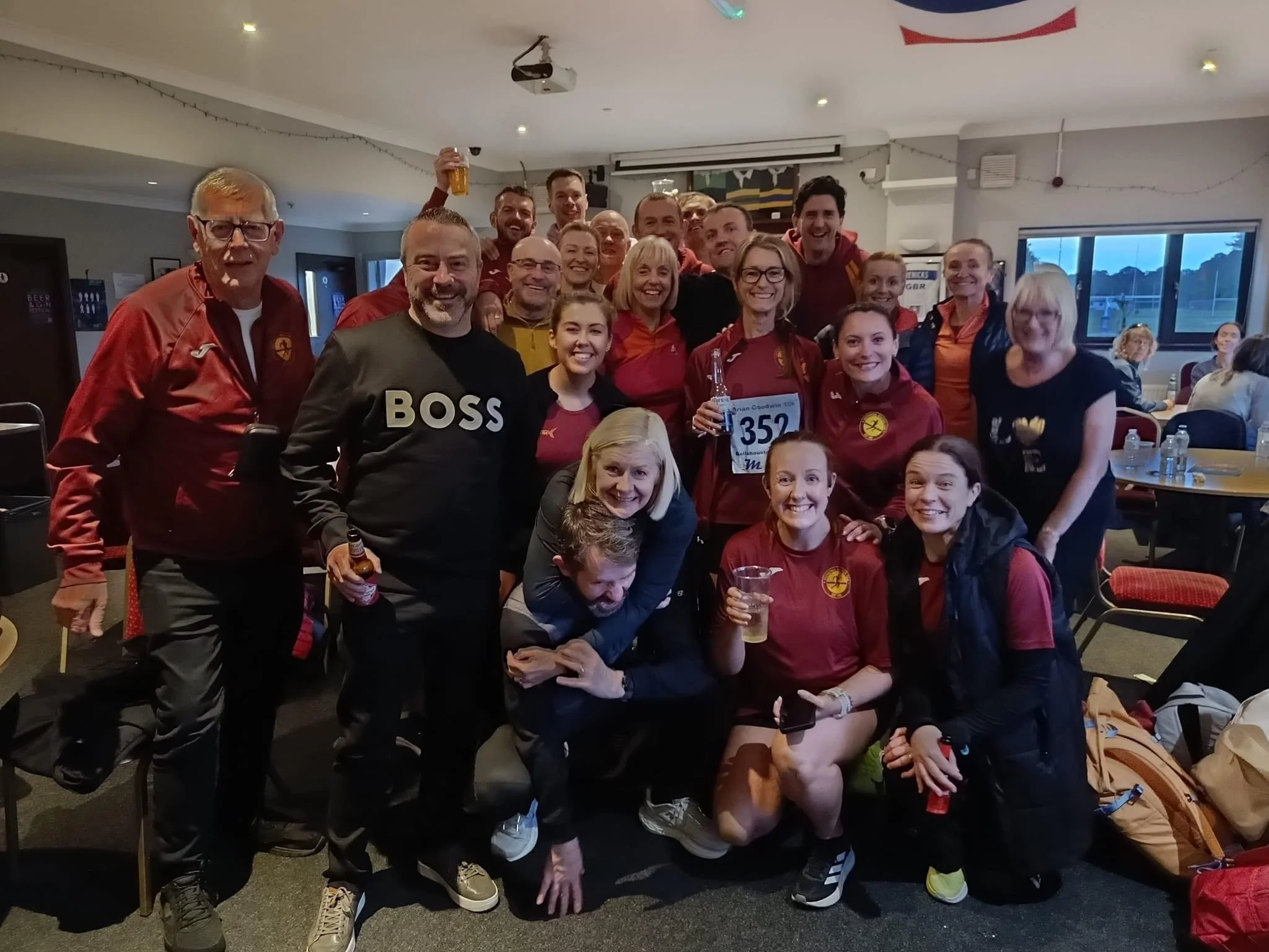 Group of people celebrating indoors, some wearing sports attire and holding drinks, smiling and posing for the photo.