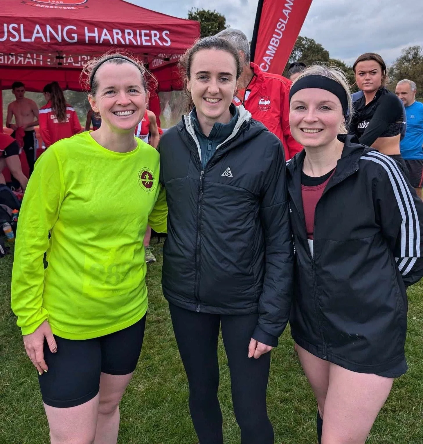Three women smiling outdoors at a running event with a red tent and other participants in the background.