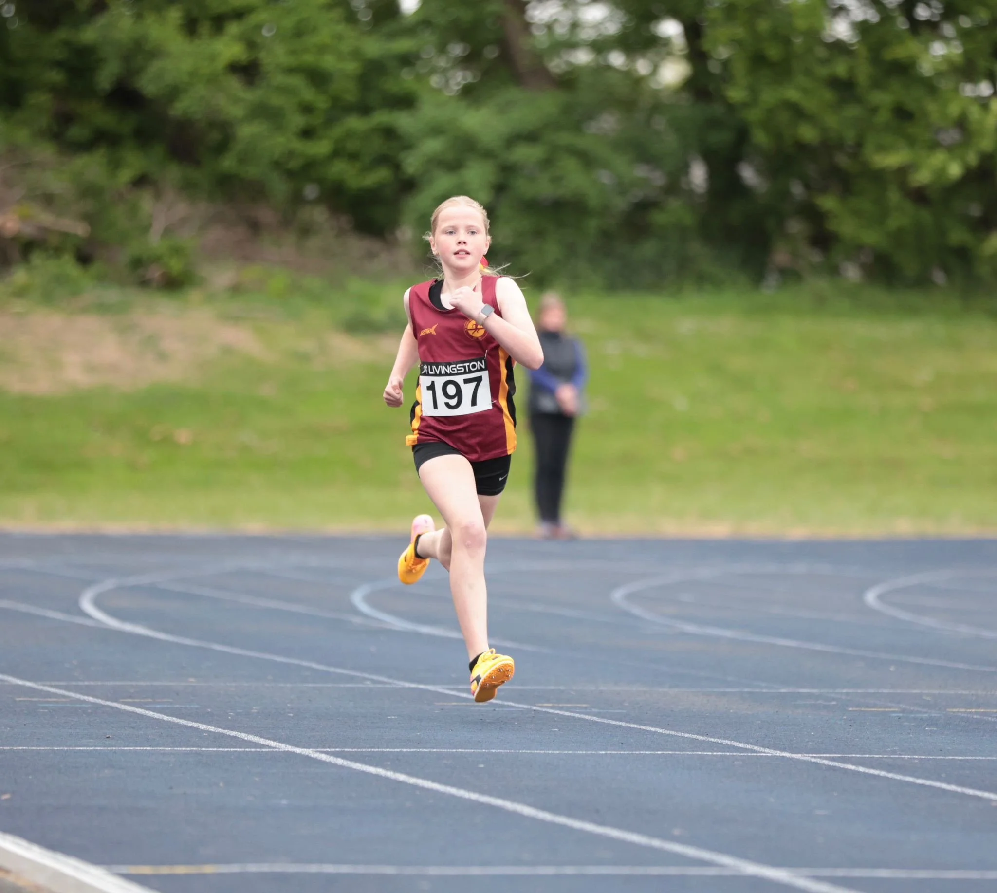 Young girl running on a track during a race, wearing a maroon sports uniform with the number 197, and a person in the background watching.