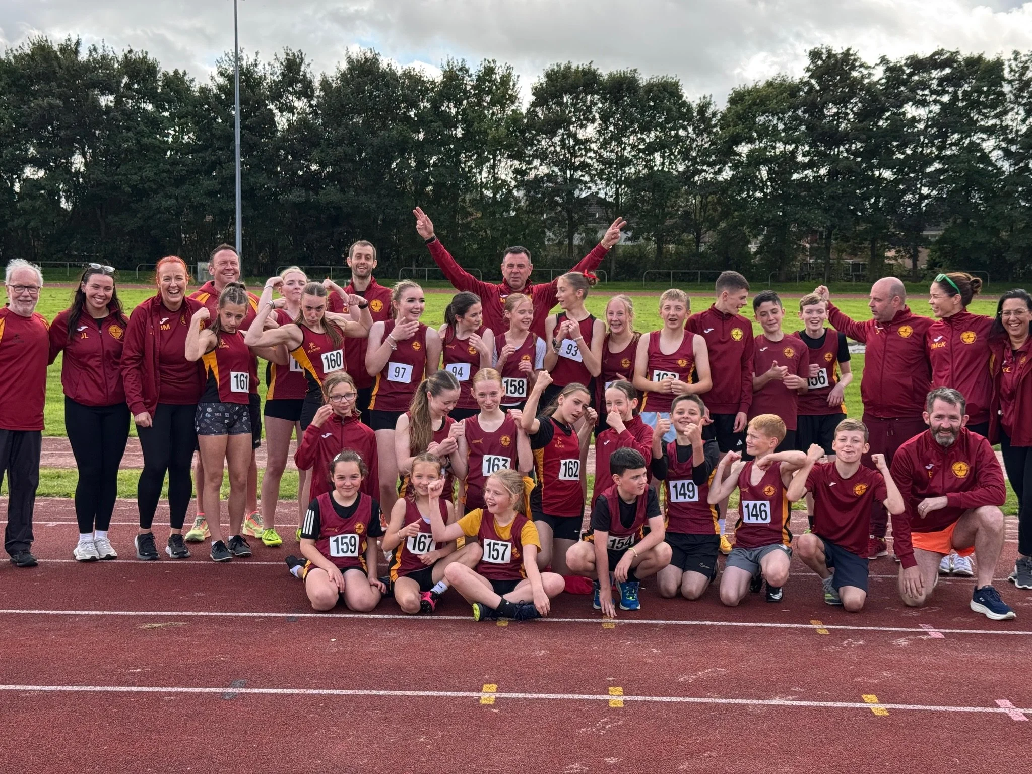 A group of young athletes and their coaches posing on a track field after a race, all wearing maroon and yellow sports uniforms, with some making victory gestures.