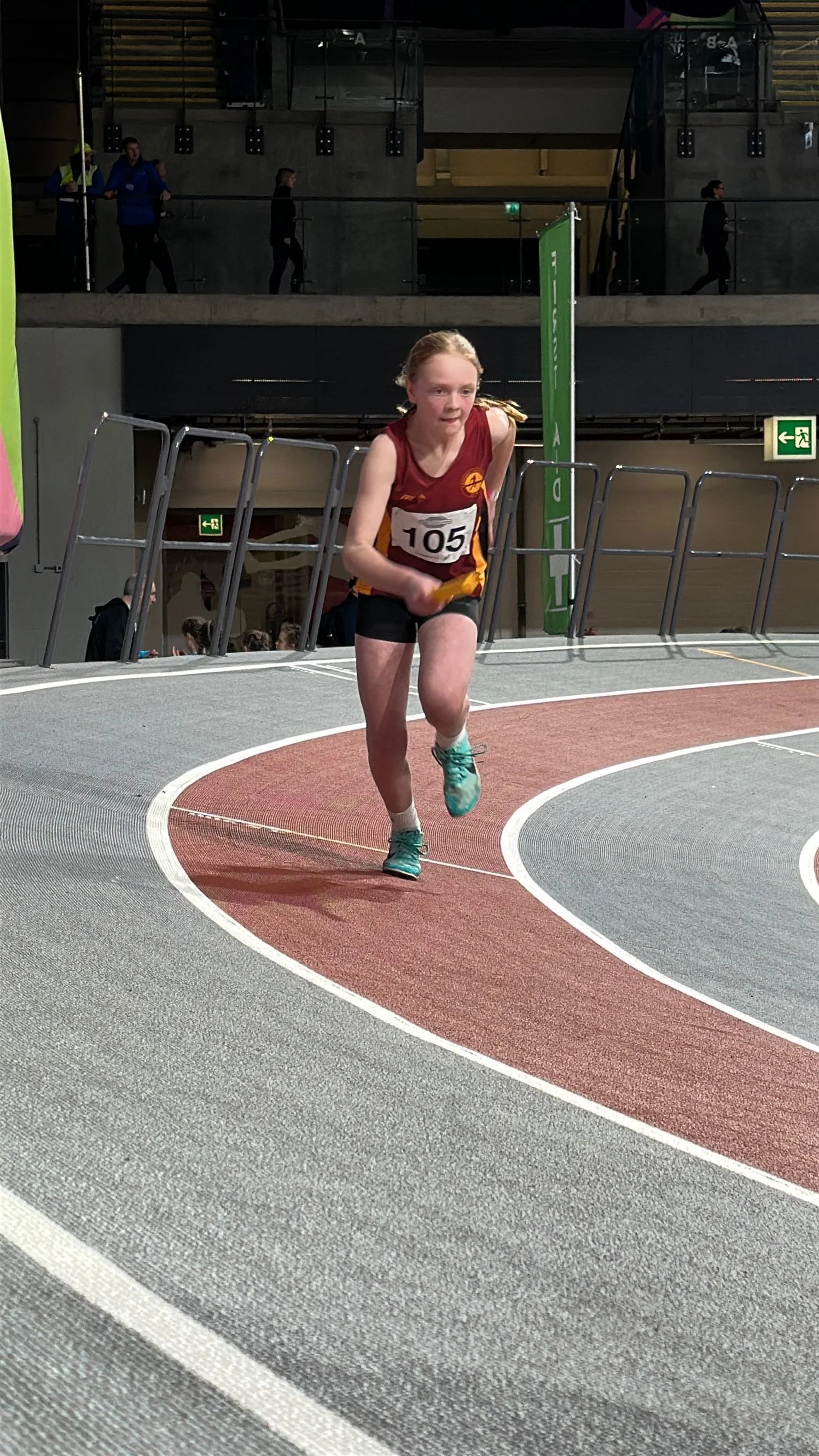 Young girl in a maroon and yellow athletic uniform running on an indoor track with a focused expression, wearing bib number 105, with spectators and other athletes in the background.