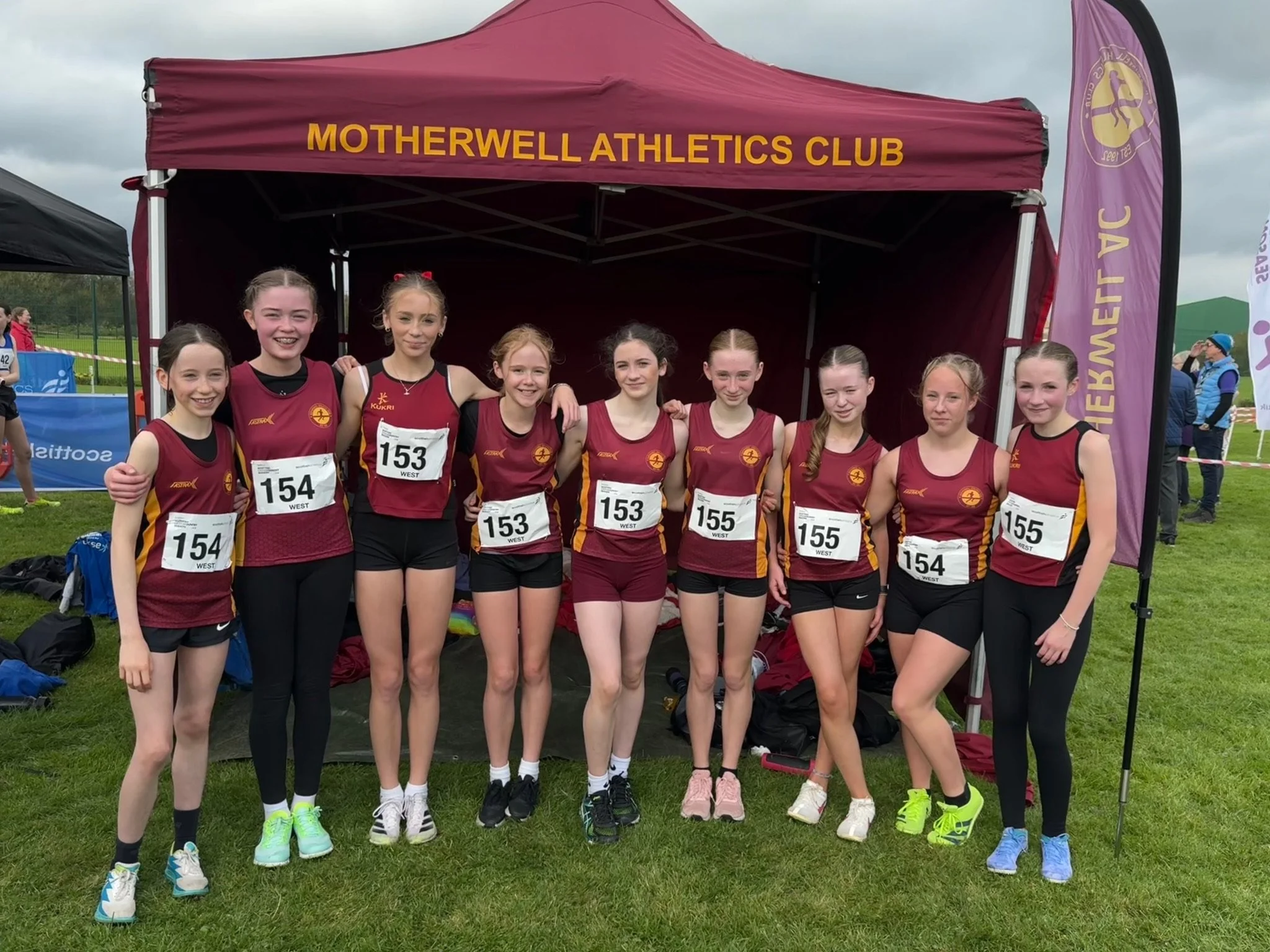 Group of young female athletes in maroon and yellow sports uniforms standing together under a maroon tent that reads 'Motherwell Athletics Club,' smiling and posing for a photo at an outdoor track event.