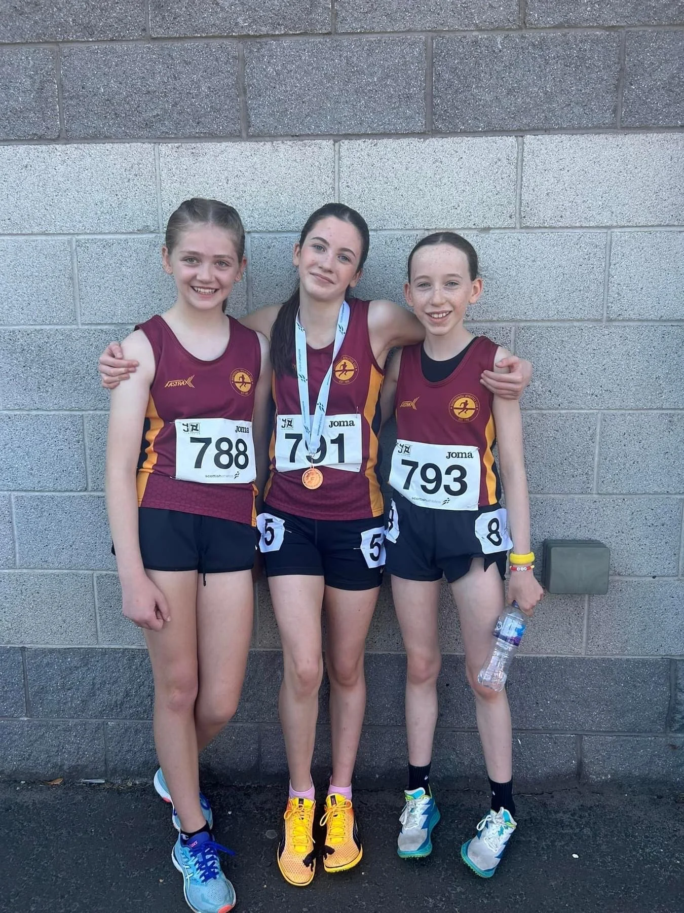Three young female runners in maroon and yellow uniforms with race numbers 788, 791, and 793, standing together with arms around each other after a race. One is wearing a medal around her neck and holding a water bottle, smiling in front of a gray br