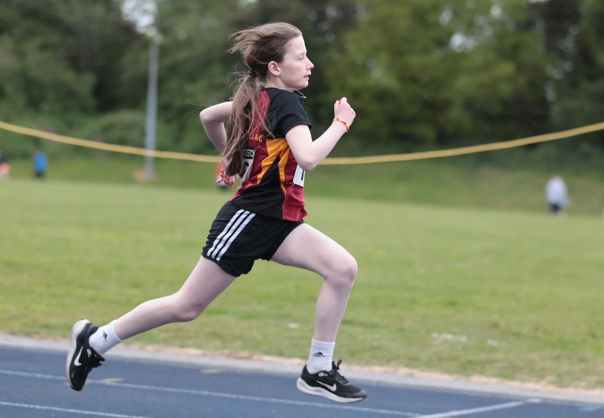A young girl running on a track during a race, wearing a sports uniform with black shorts, a black and red shirt, and running shoes, with grassy field and trees in the background.