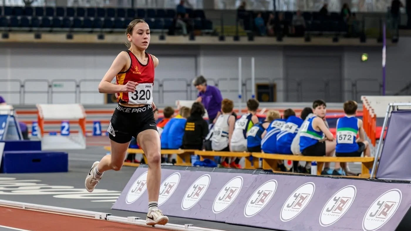 A young female athlete in red and black sports attire running in an indoor track and field stadium.