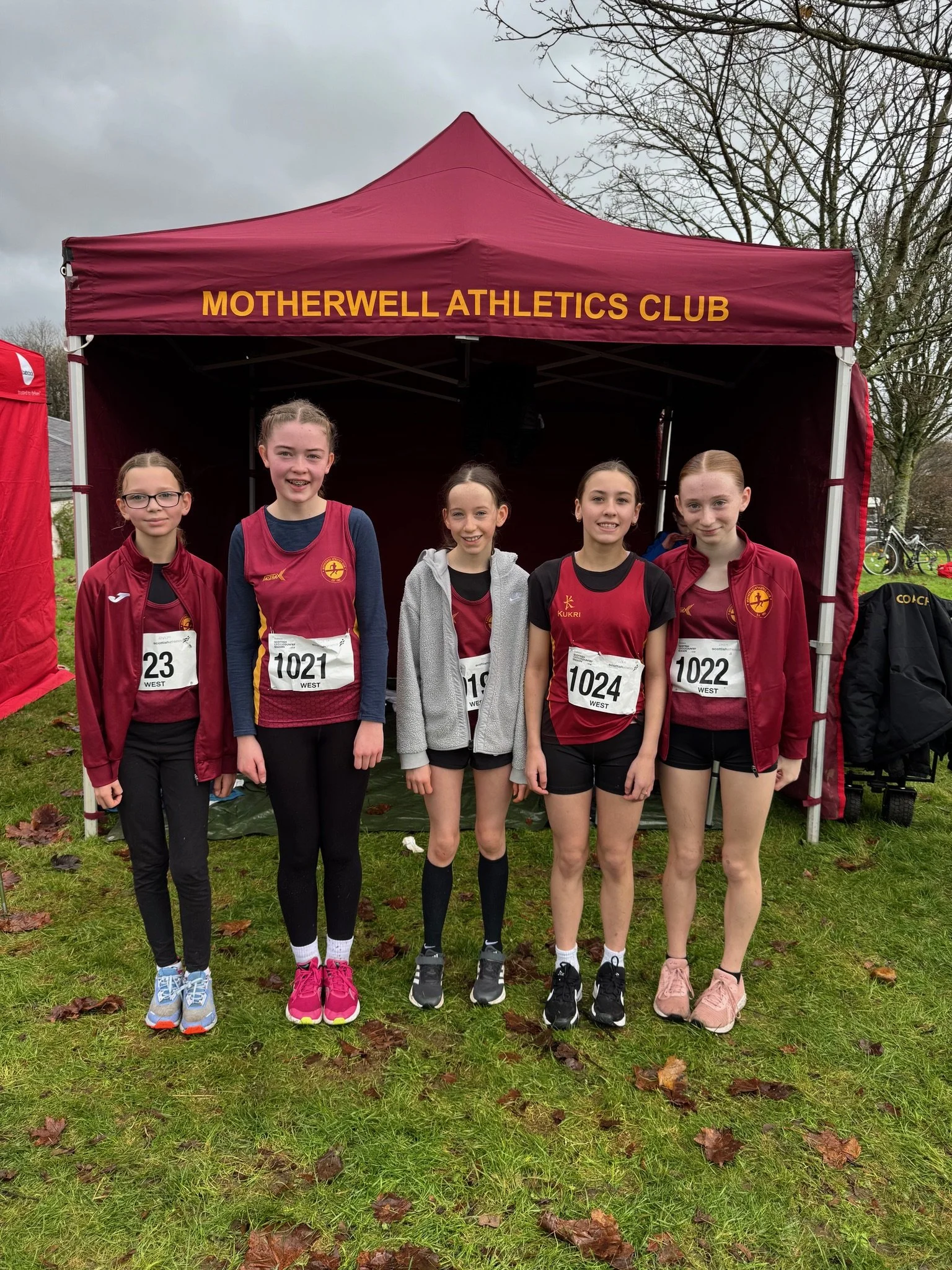 Group of five young female athletes standing in front of a maroon tent with yellow text that reads 'Motherwell Athletics Club'. They are dressed in sports attire, some with race bibs, and appear to be at an outdoor track or park on a cloudy day.