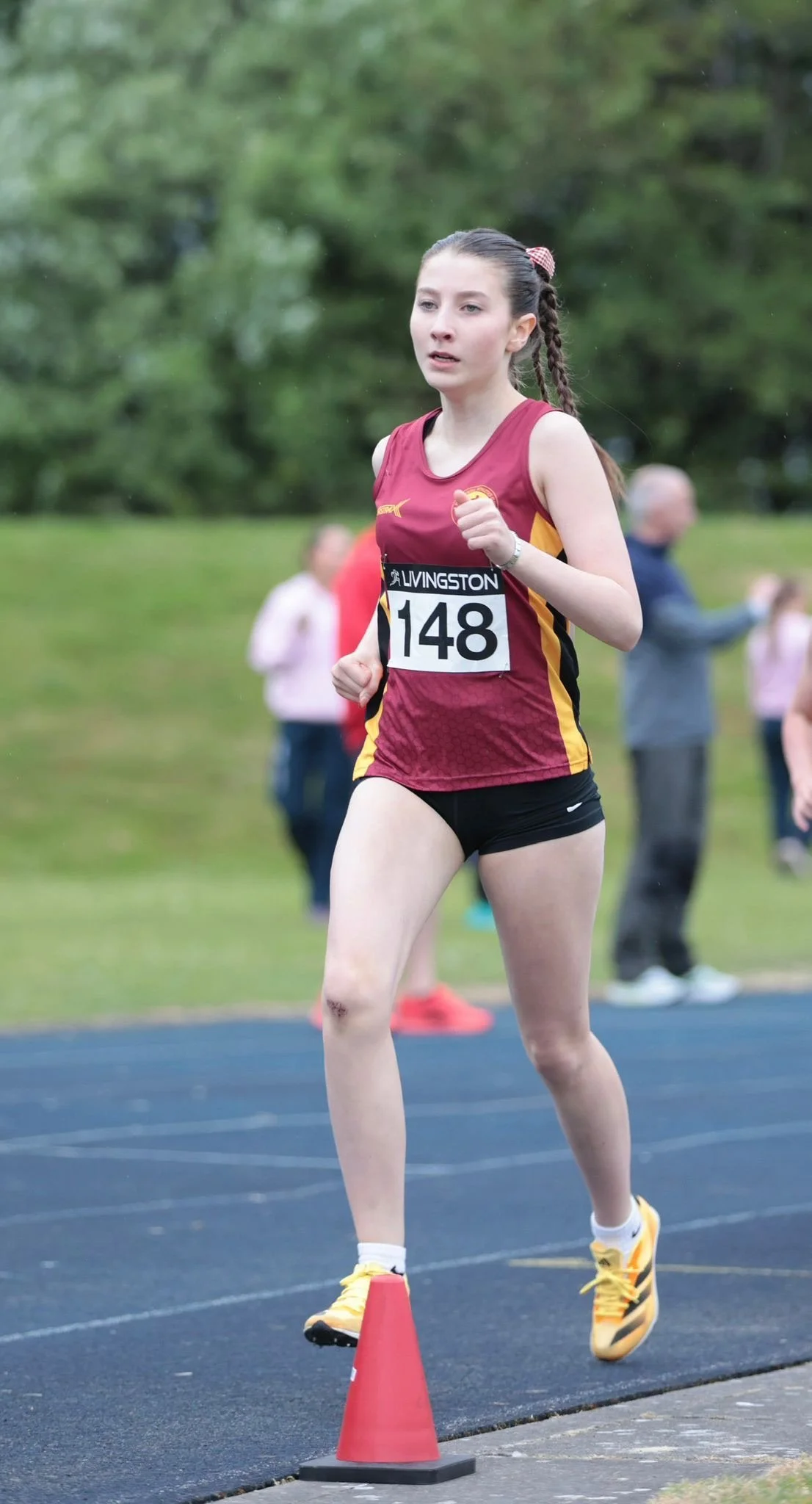 A young female athlete running on a track during a race, wearing a maroon and yellow athletic uniform with the number 148. She is navigating around a red cone with her hair in braids and a focused expression.