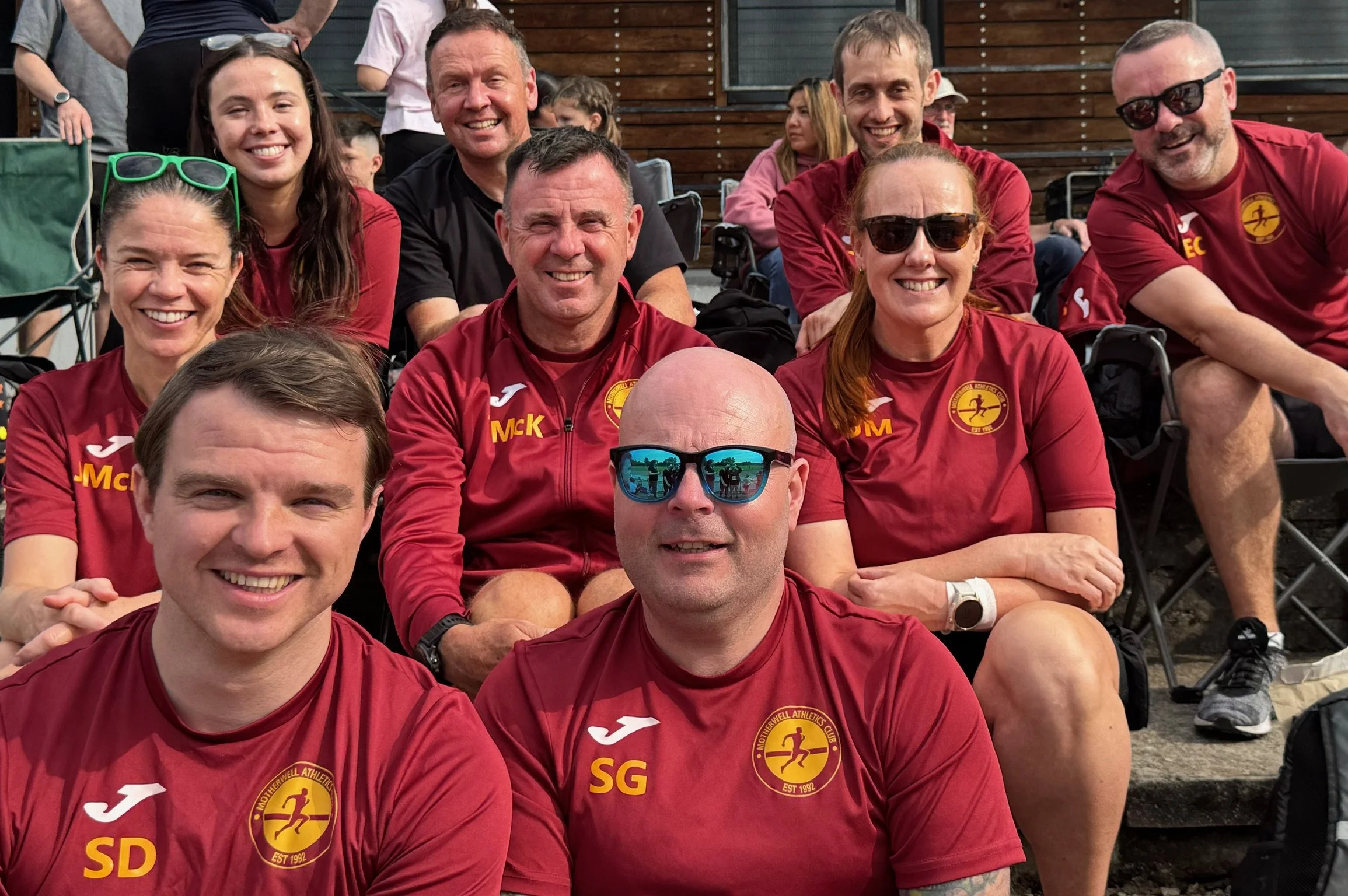 Group of ten smiling people, wearing matching red athletic shirts with the logo and initials of Motherwell Athletics Club, posing outdoors on stadium steps.
