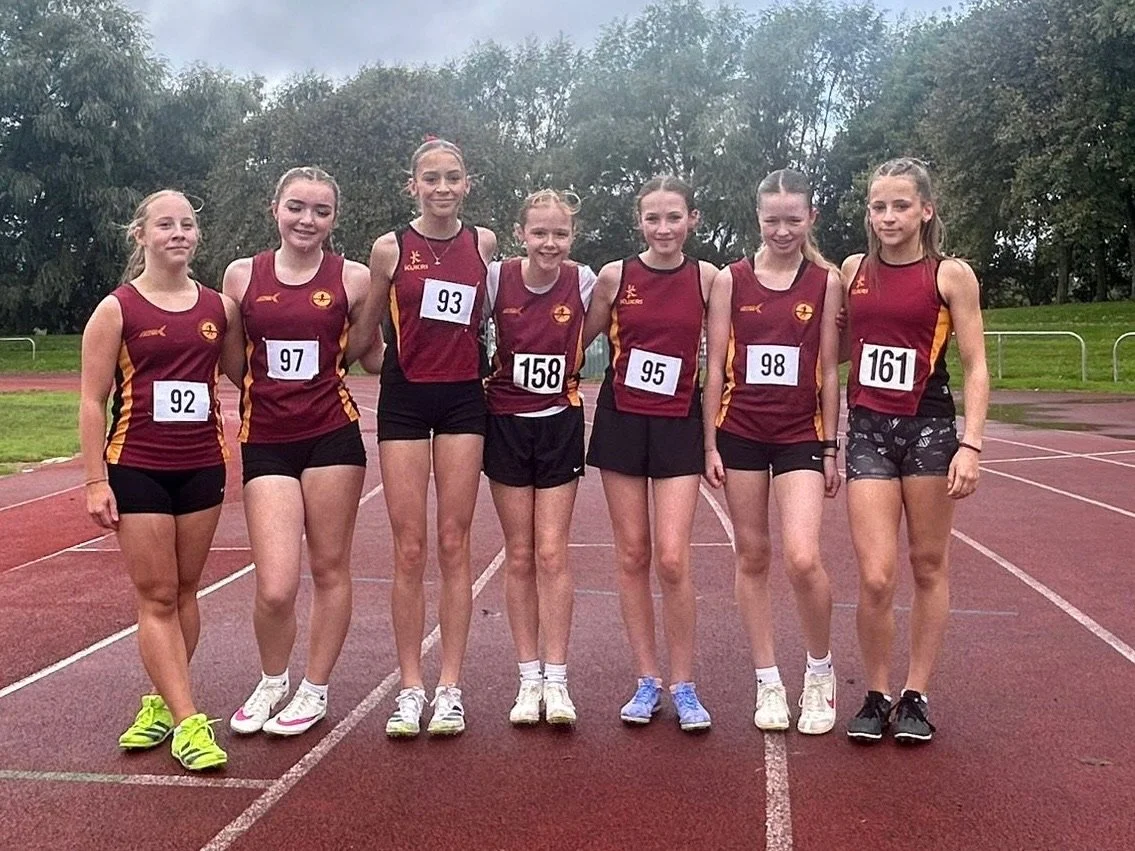 Seven young female athletes standing on a running track, posing for a group photo before a race. They are wearing maroon and yellow sports uniforms with race numbers on their chests.