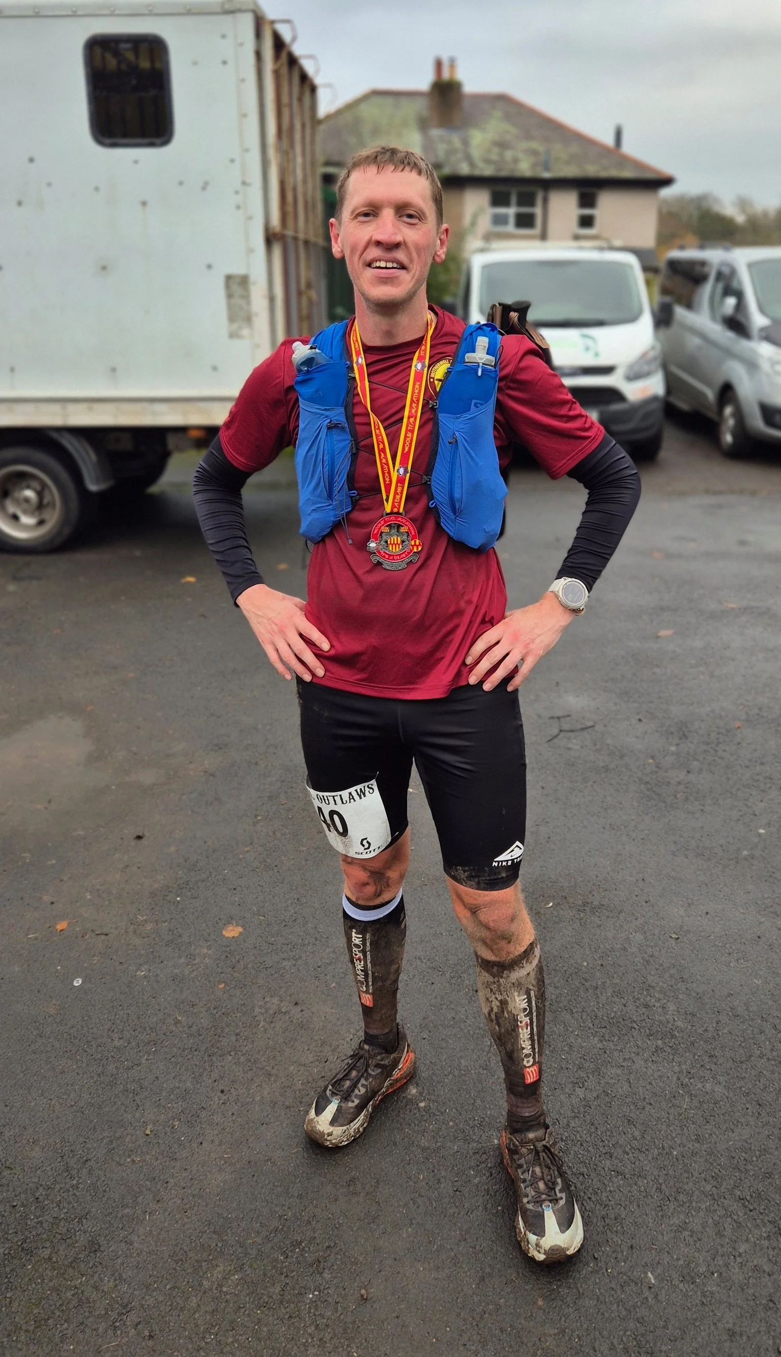 A man in running gear, wearing a red shirt, black shorts, and muddy trail running shoes, stands outdoors with hands on hips, smiling after a race, with a gold medal around his neck.