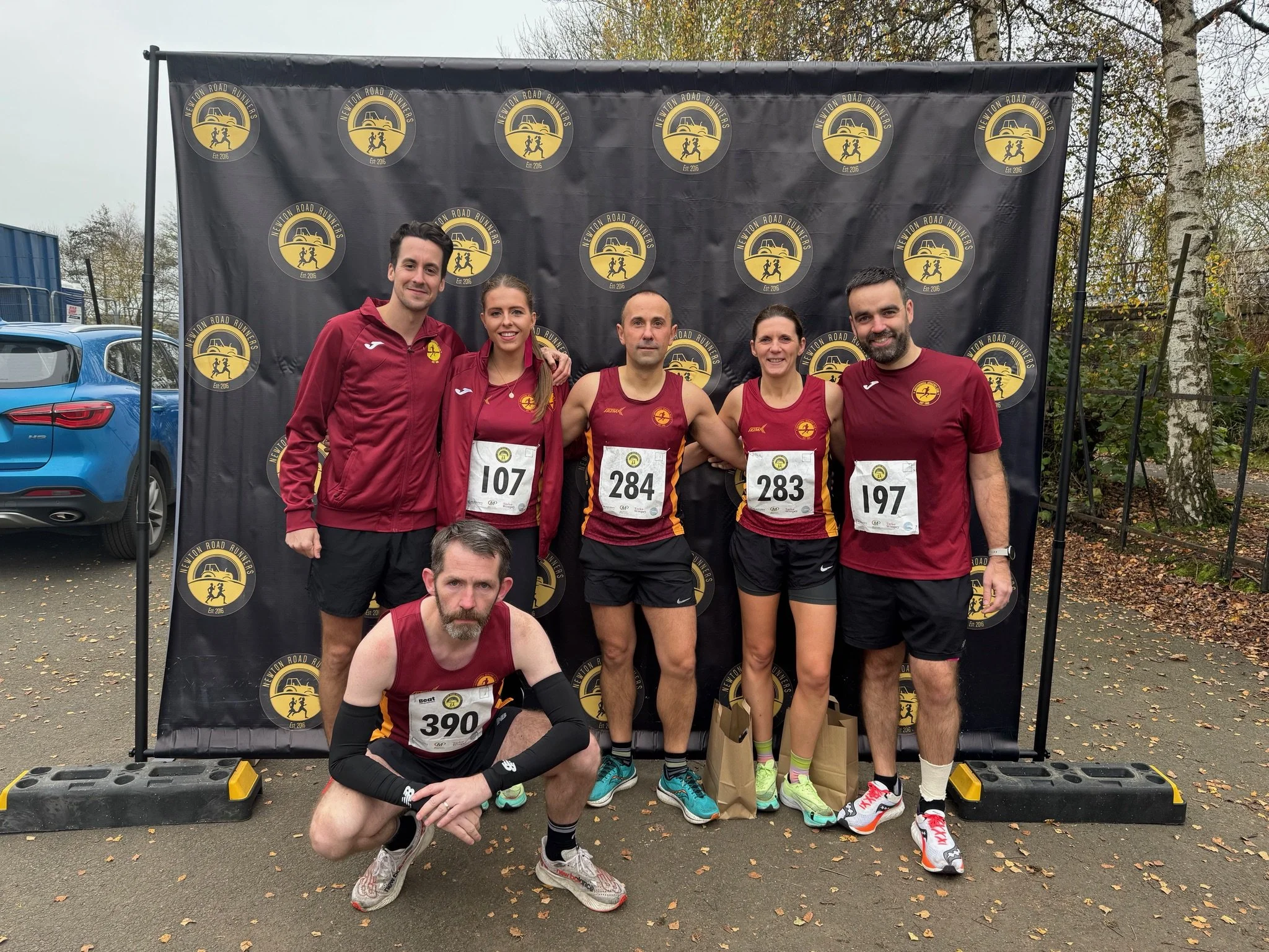 Six runners in maroon and black athletic gear posing in front of a backdrop with a circular logo of a yellow car and running figures, at a race event. Some are standing, one is kneeling.