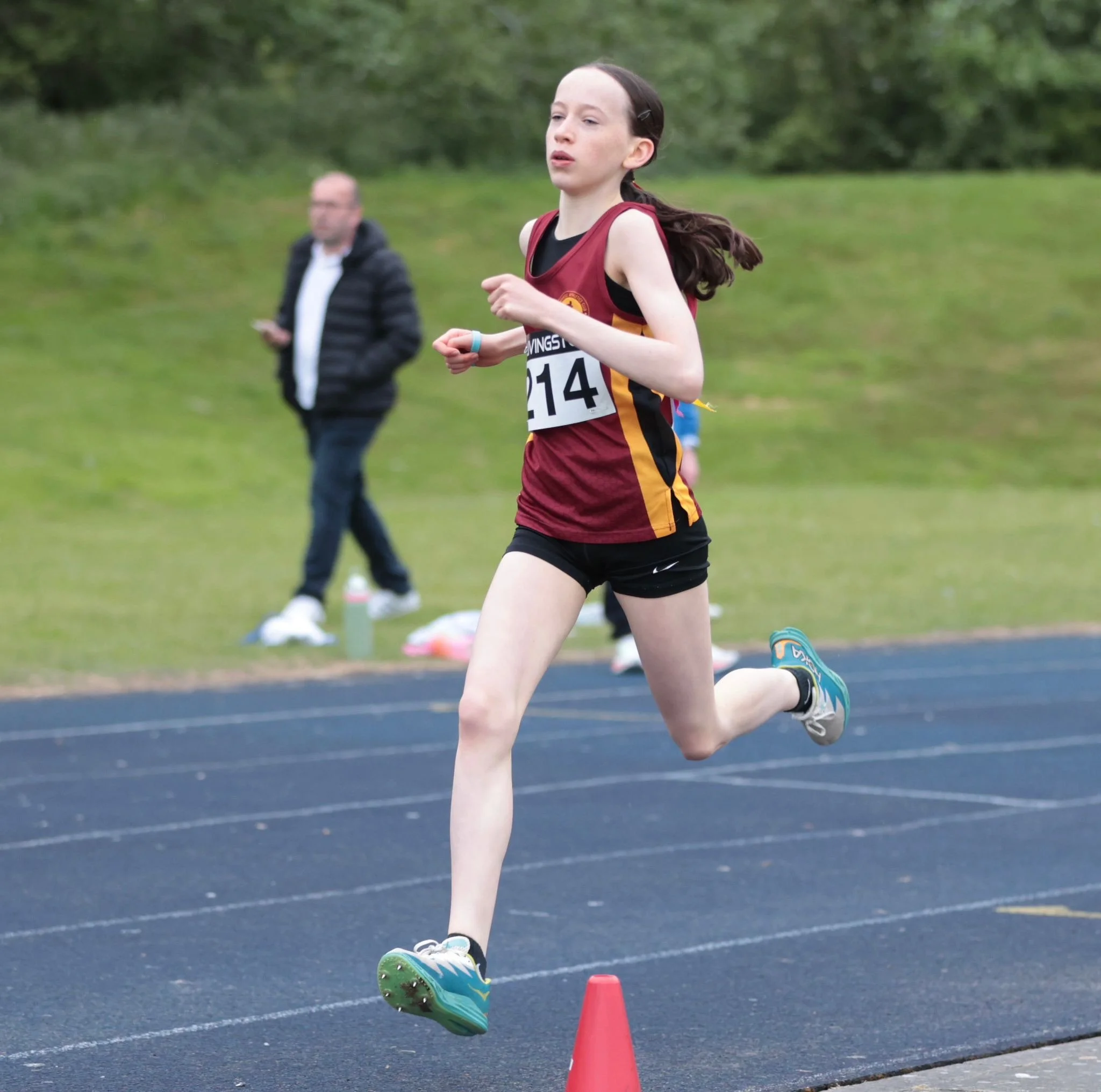 A young female athlete wearing a maroon and gold running uniform with the number 214, running on a track during a race, with a man in the background on a grassy field.