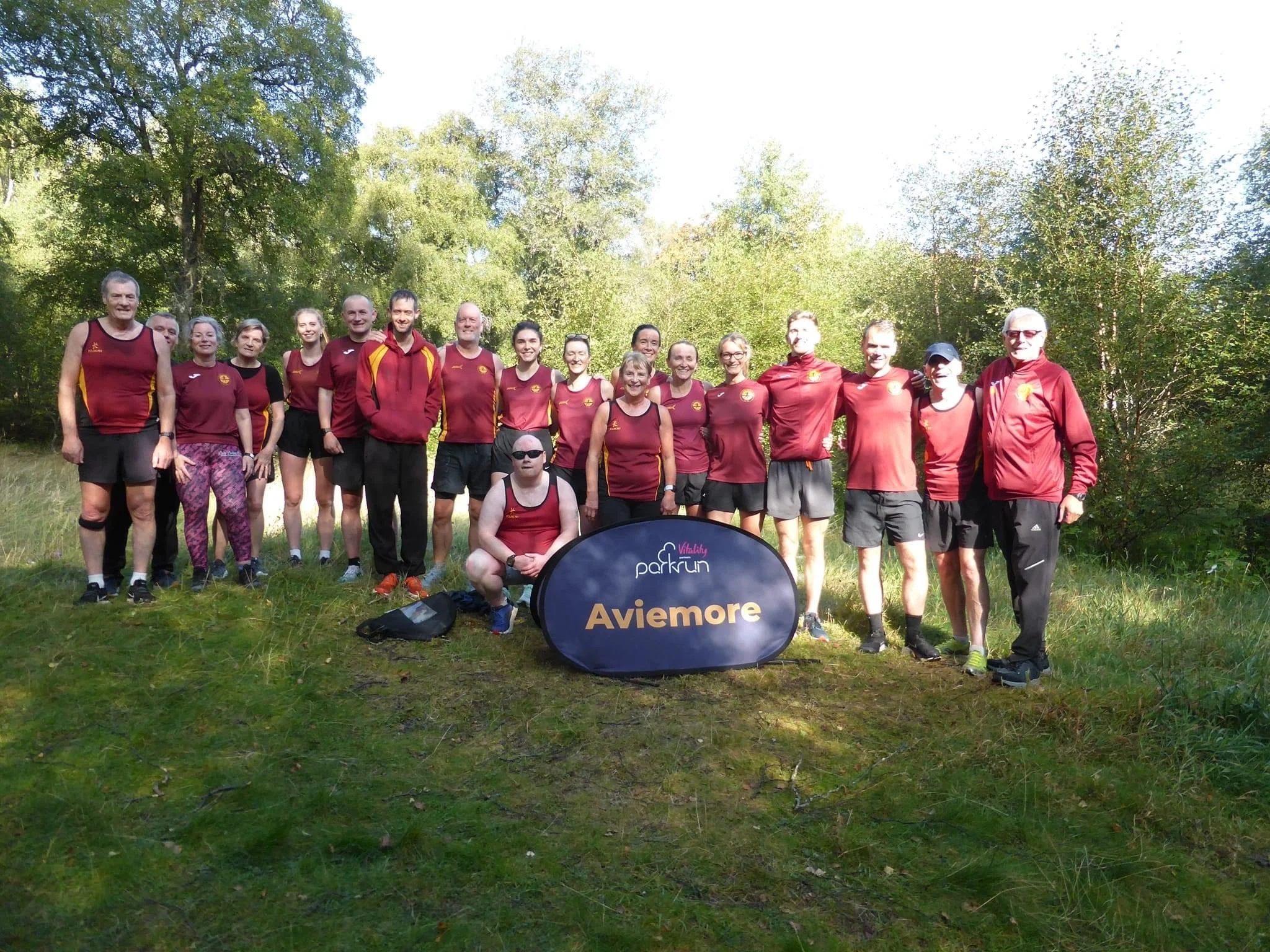 Group of people in athletic clothing standing outdoors on grass with trees in the background, holding a sign that reads 'Aviemore'.