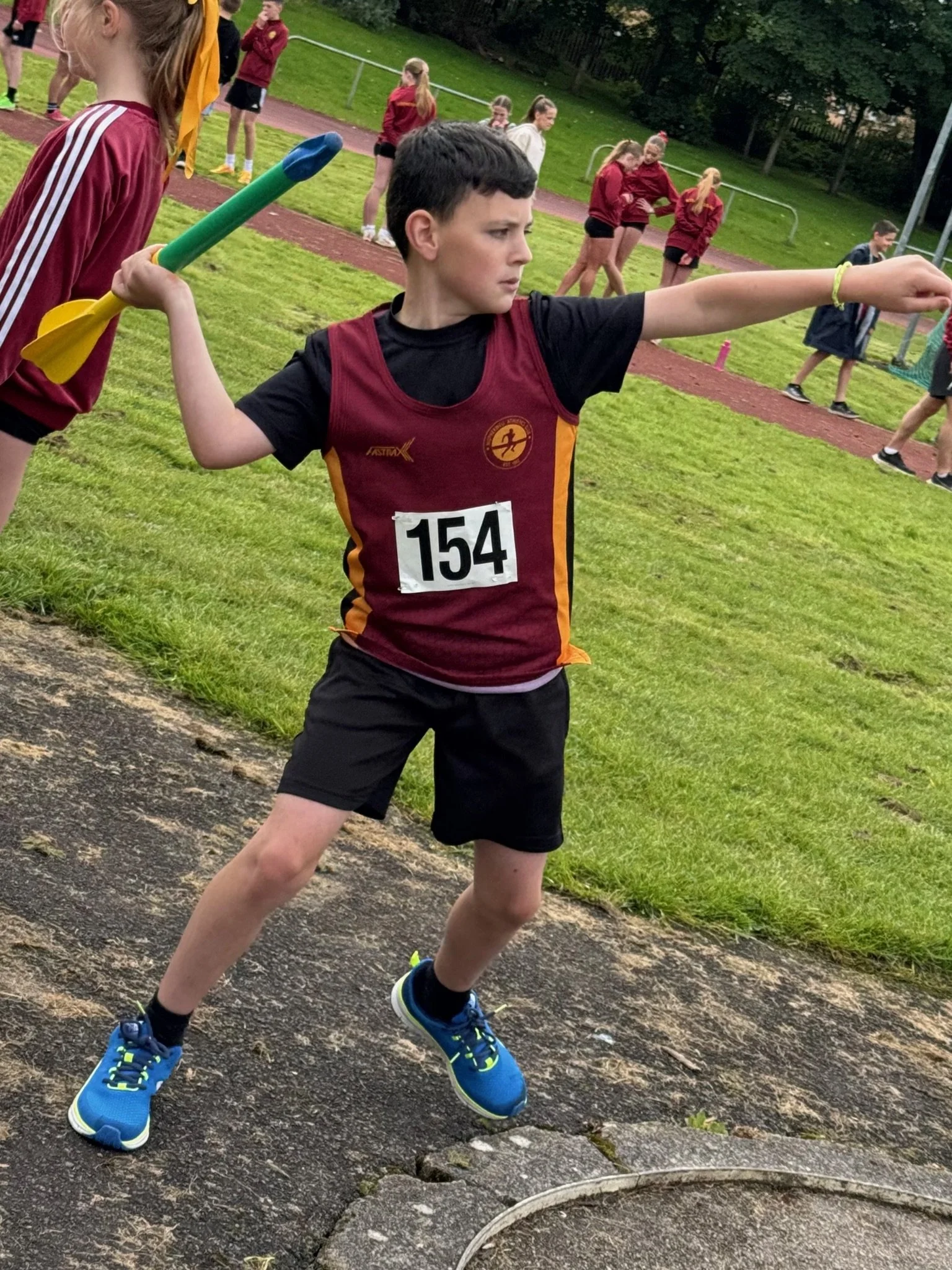 A young boy participating in a track and field event, holding a javelin in his right hand, standing on a track with a grassy field and other children in the background.