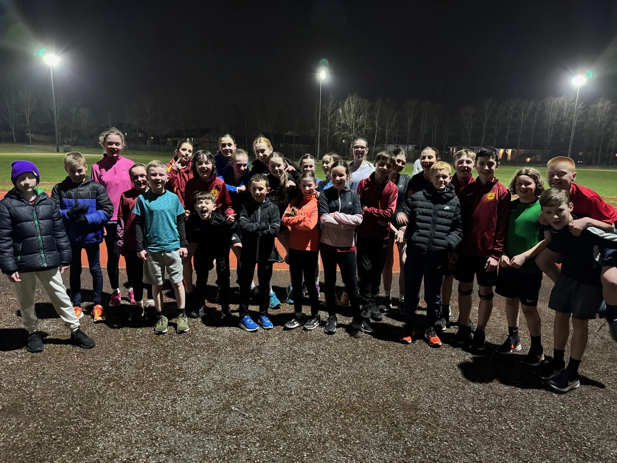 Group of children and teenagers posing for a photo on a running track at night under bright stadium lights.
