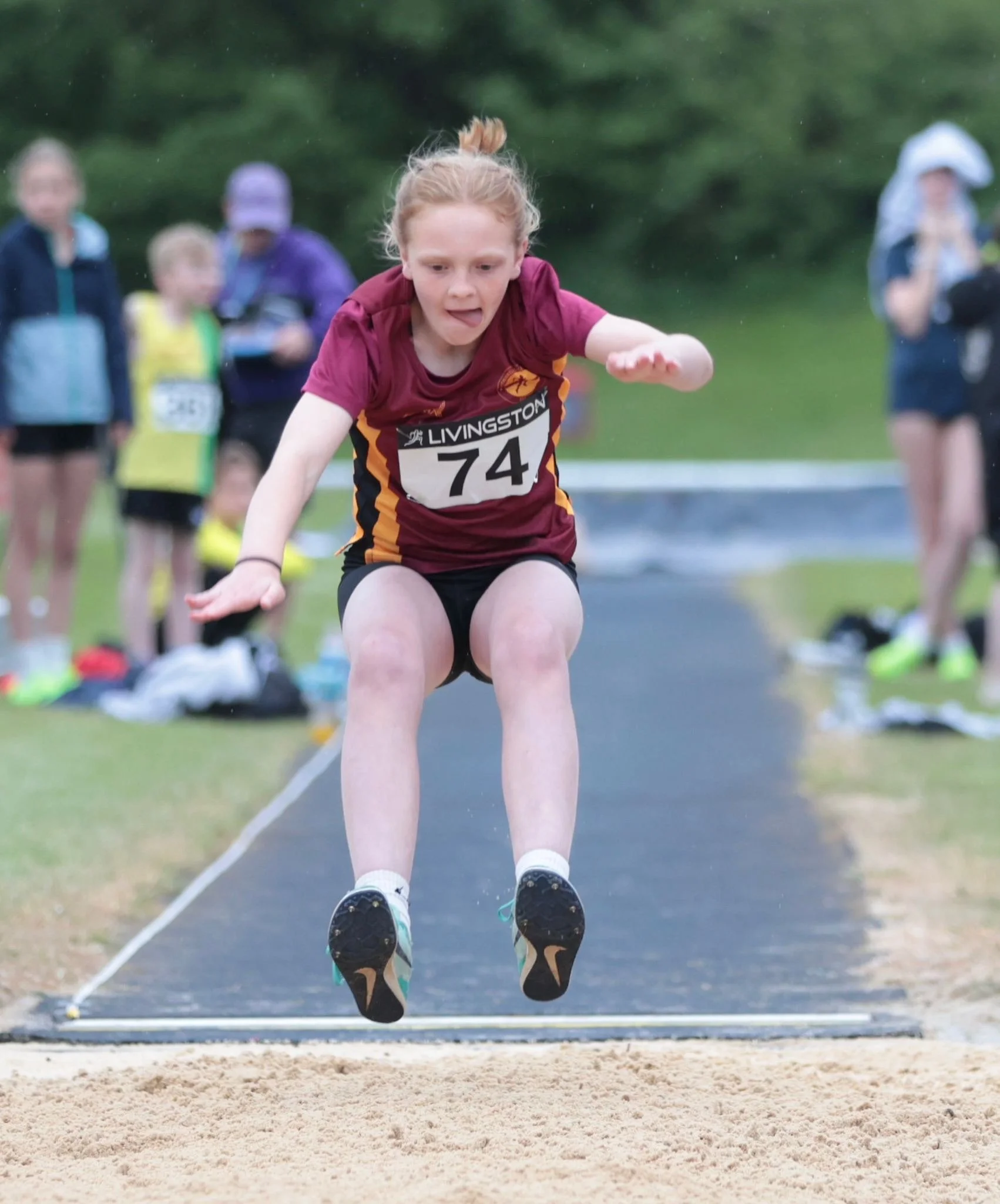 Young female athlete in maroon and gold uniform jumping into sandpit during long jump event at track and field competition with spectators in background.