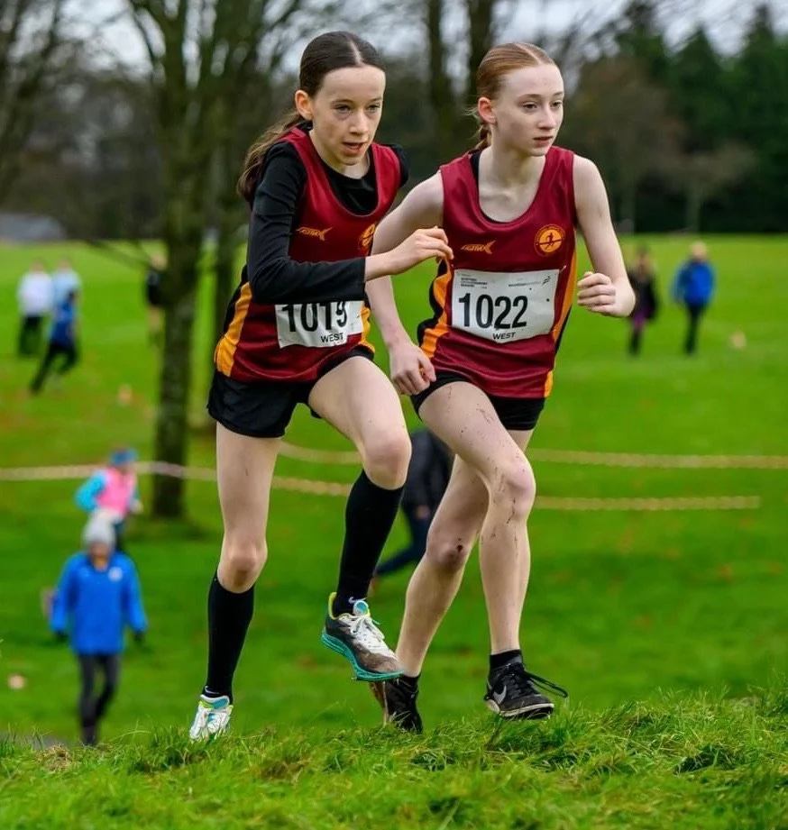Two young female cross-country runners in red and black uniforms competing on a grassy outdoor track, with spectators in the background.