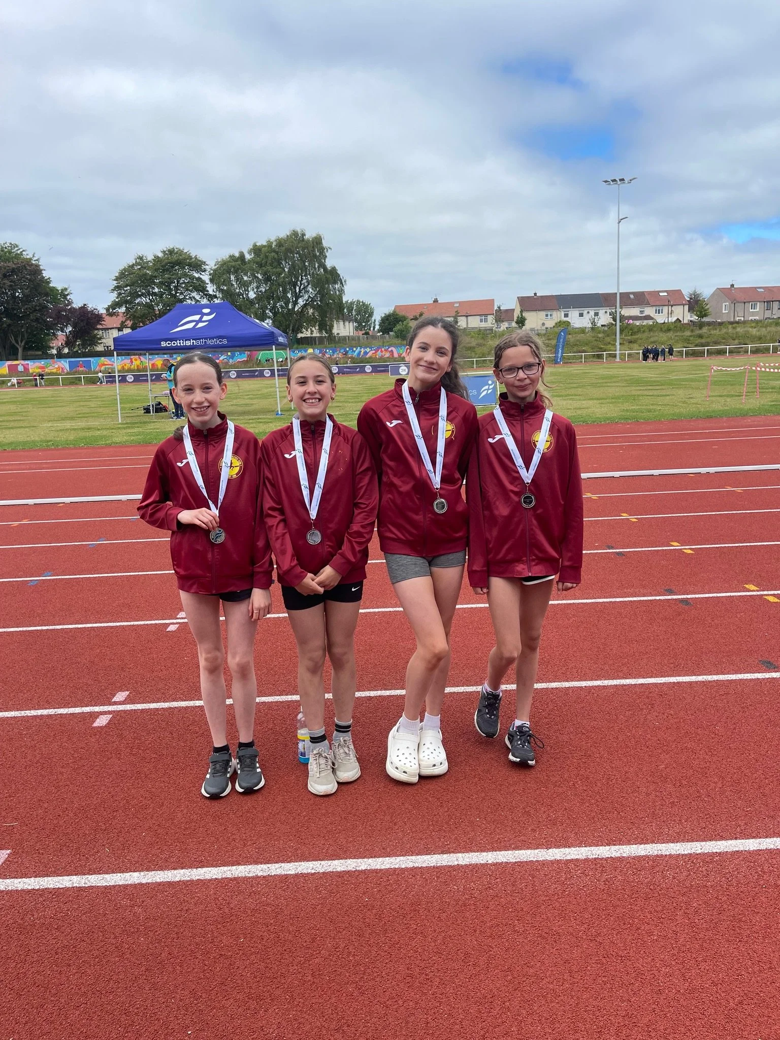 Four young girls standing on a track, wearing matching maroon jackets and silver medals around their necks, smiling for a photo after a track and field event.