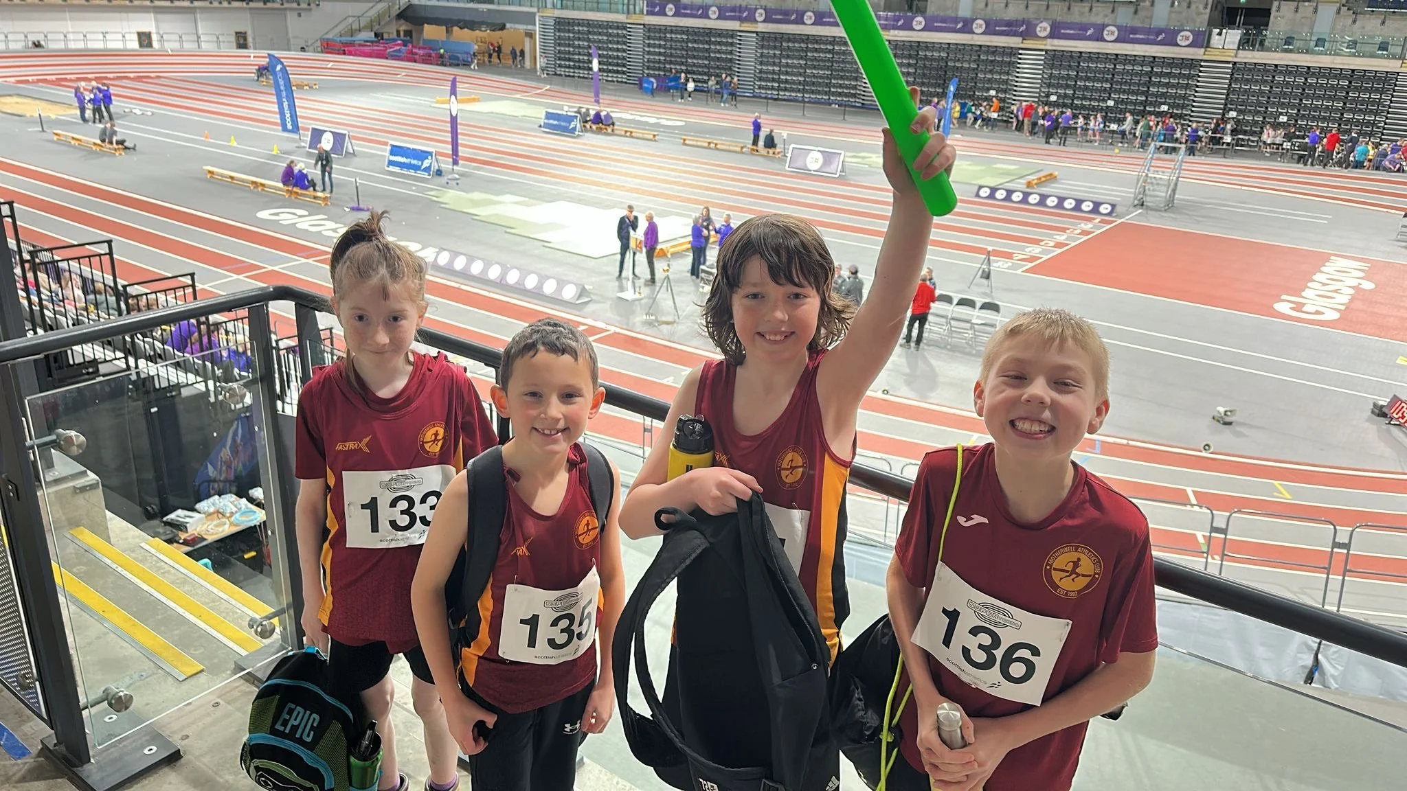 Four young children wearing red athletic uniforms with race bibs and backpacks at an indoor track and field stadium, smiling and posing for the photo.