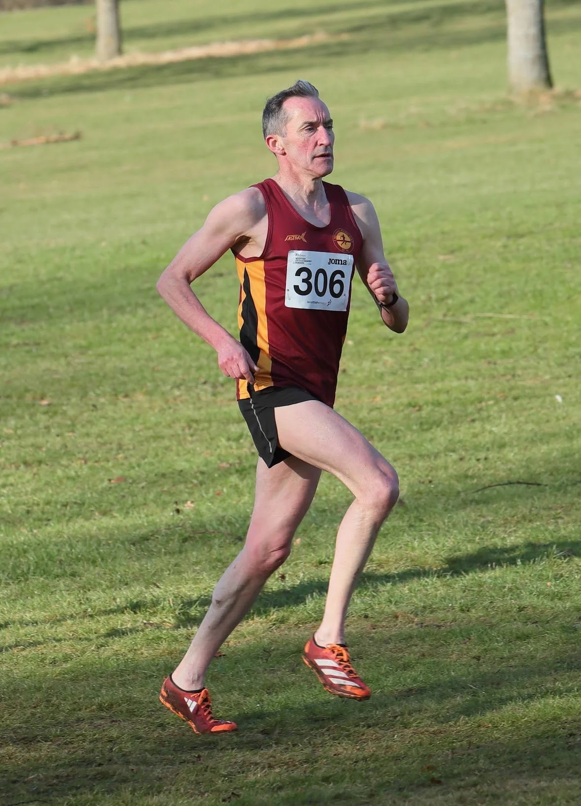 A male runner wearing a maroon and yellow athletic uniform and orange running shoes, participating in a race on a grassy field.