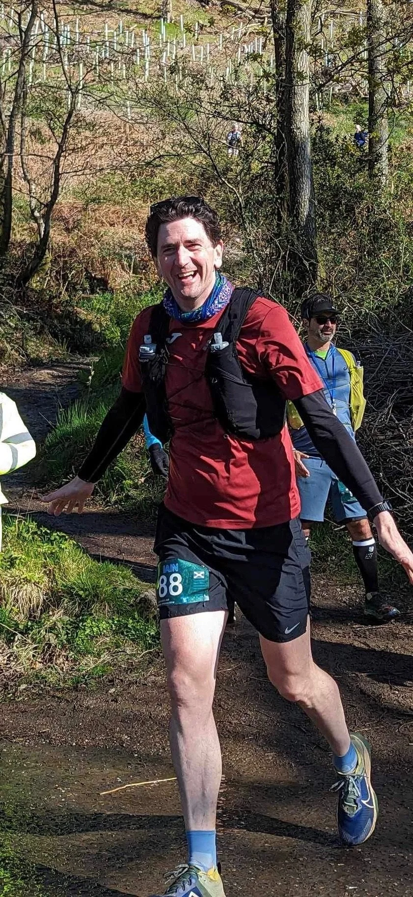 A man running on a trail in a forest, smiling, wearing a red athletic shirt, black shorts, and running gear during a race.