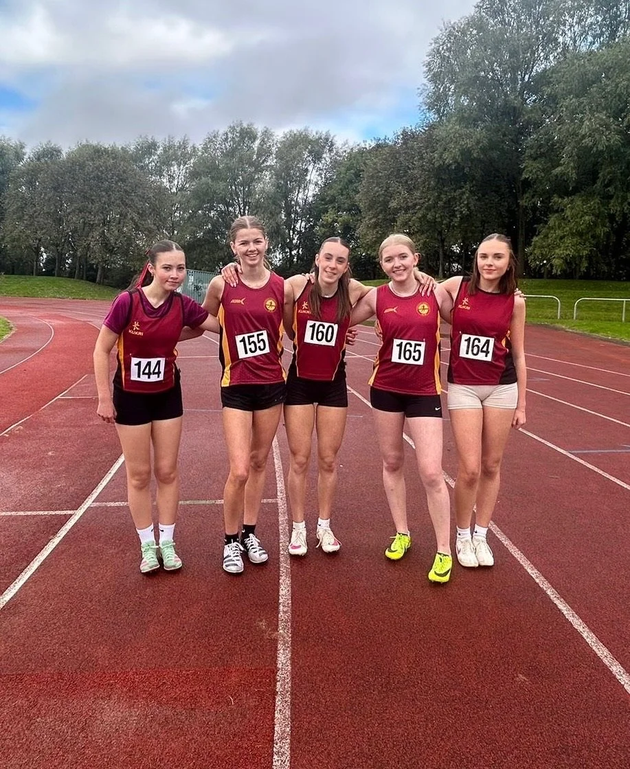 Five young girls in athletic uniforms standing on a track, smiling, with trees and cloudy sky in the background.