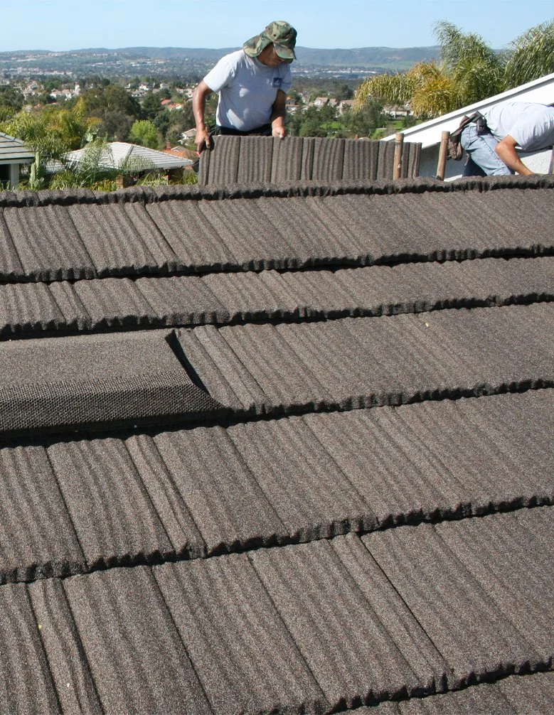 Two workers install roof tiles on a sloped roof, with a scenic view of a neighborhood and hills in the background.