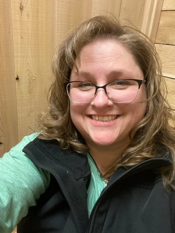 A woman with curly, light brown hair and glasses smiling at the camera, wearing a green top and a black vest, in front of a wooden wall.