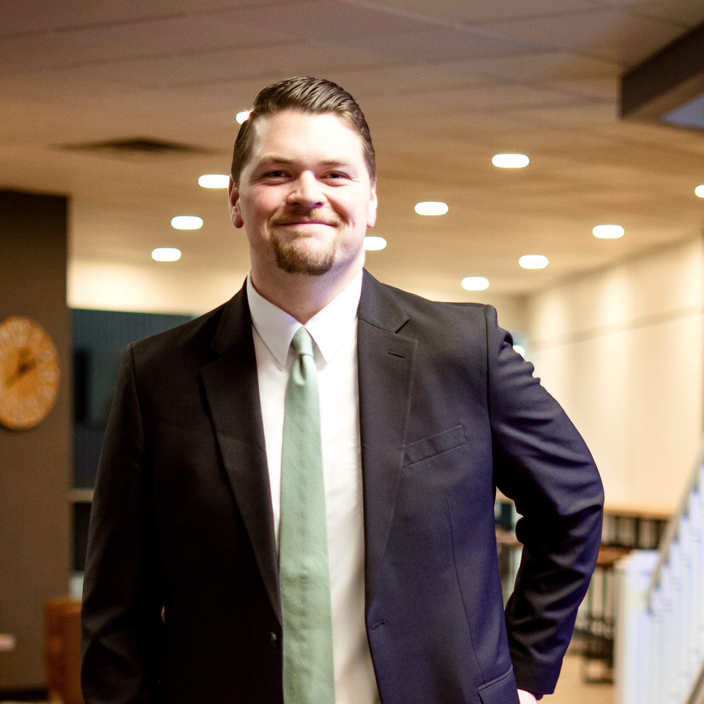 A young man with brown hair, beard, and mustache wearing a dark suit, white shirt, and light green tie, smiling in an indoor setting with warm lighting.