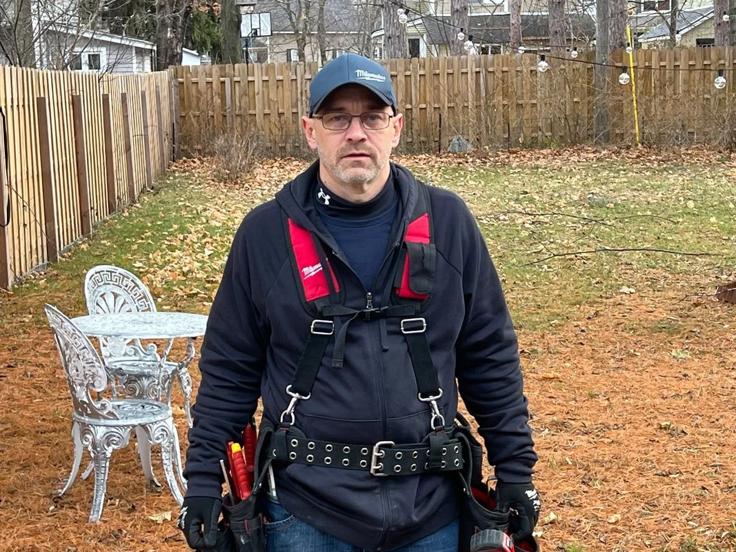 Man with safety harness standing in backyard with leaves, wooden fence, white metal garden furniture, and string lights.