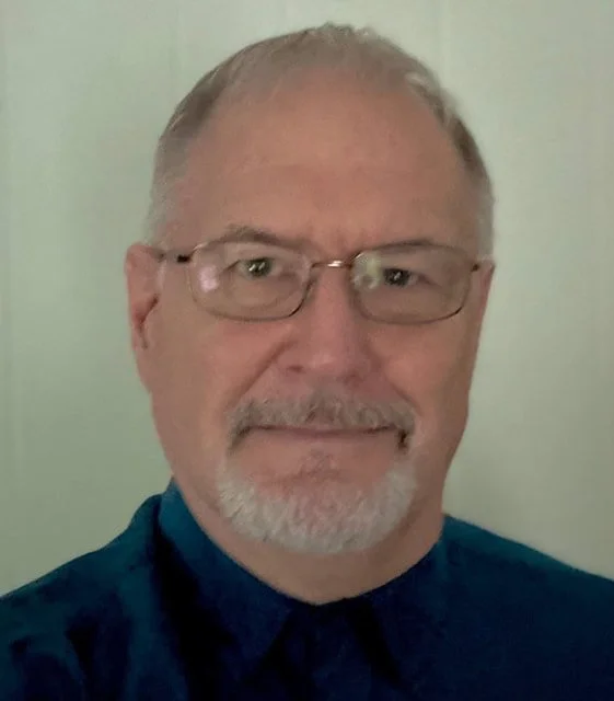 A middle-aged man with glasses, short gray hair, and a beard, wearing a dark collared shirt, standing in front of a plain light-colored wall.